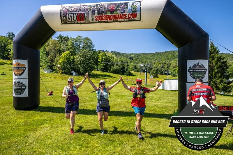 Four people celebrating under an inflatable arch at a trail race event, with a grassy field, trees, and hills in the background.