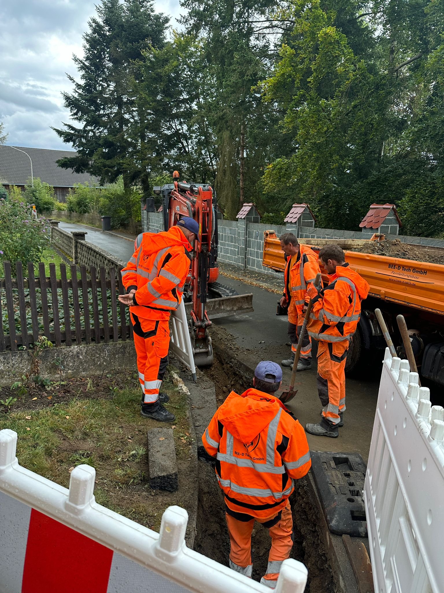Bauarbeiter in orange Warnschutzkleidung arbeiten an einer Straßensperre auf einer Nebenstraße, mit Bäumen und Häuser im Hintergrund.