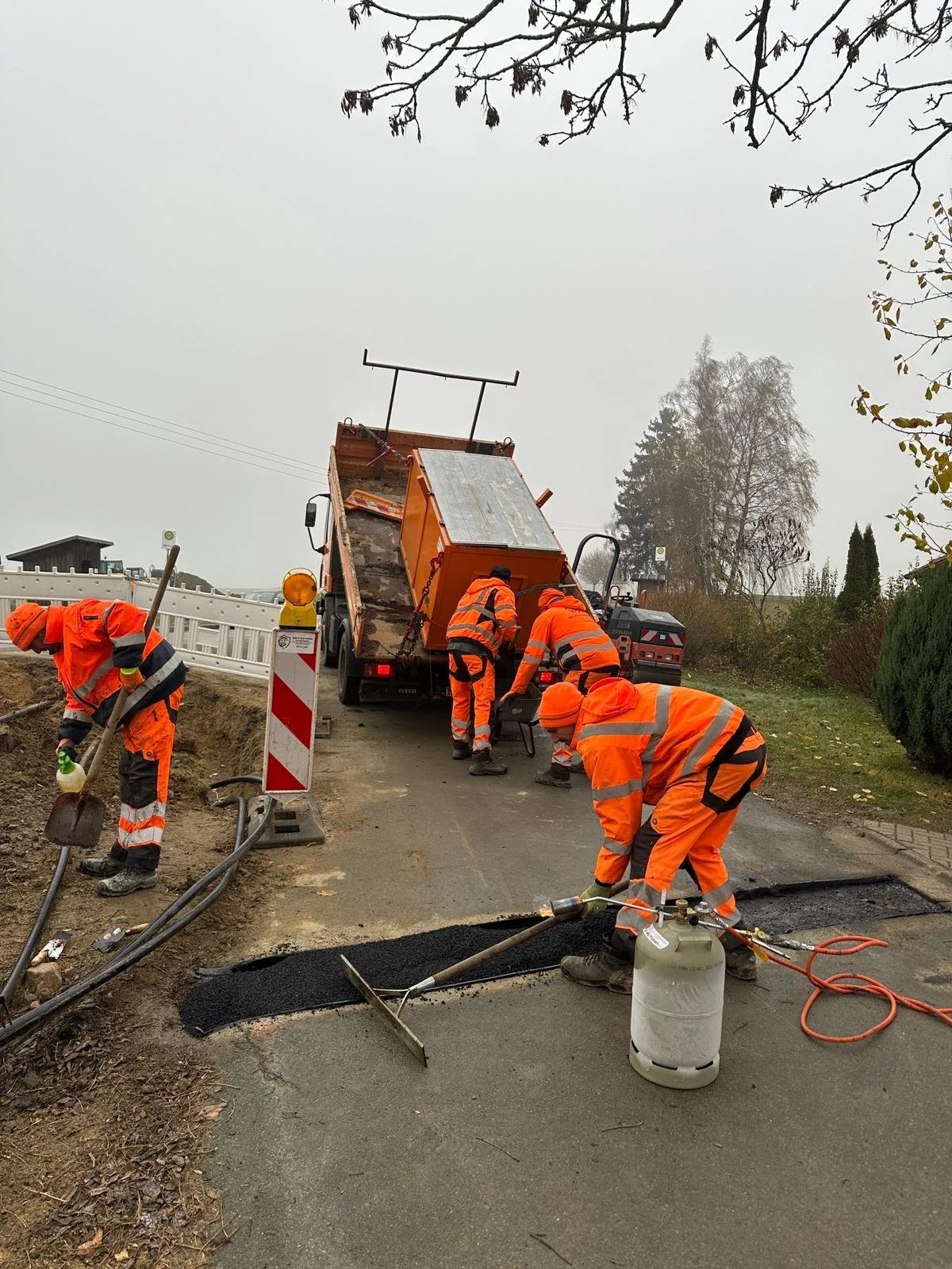 Straßenbauarbeiter in orangenen Sicherheitskleidung arbeiten an der Asphaltierung einer Straße, mit einem Fahrzeug im Hintergrund und einem Baum im Himmel.