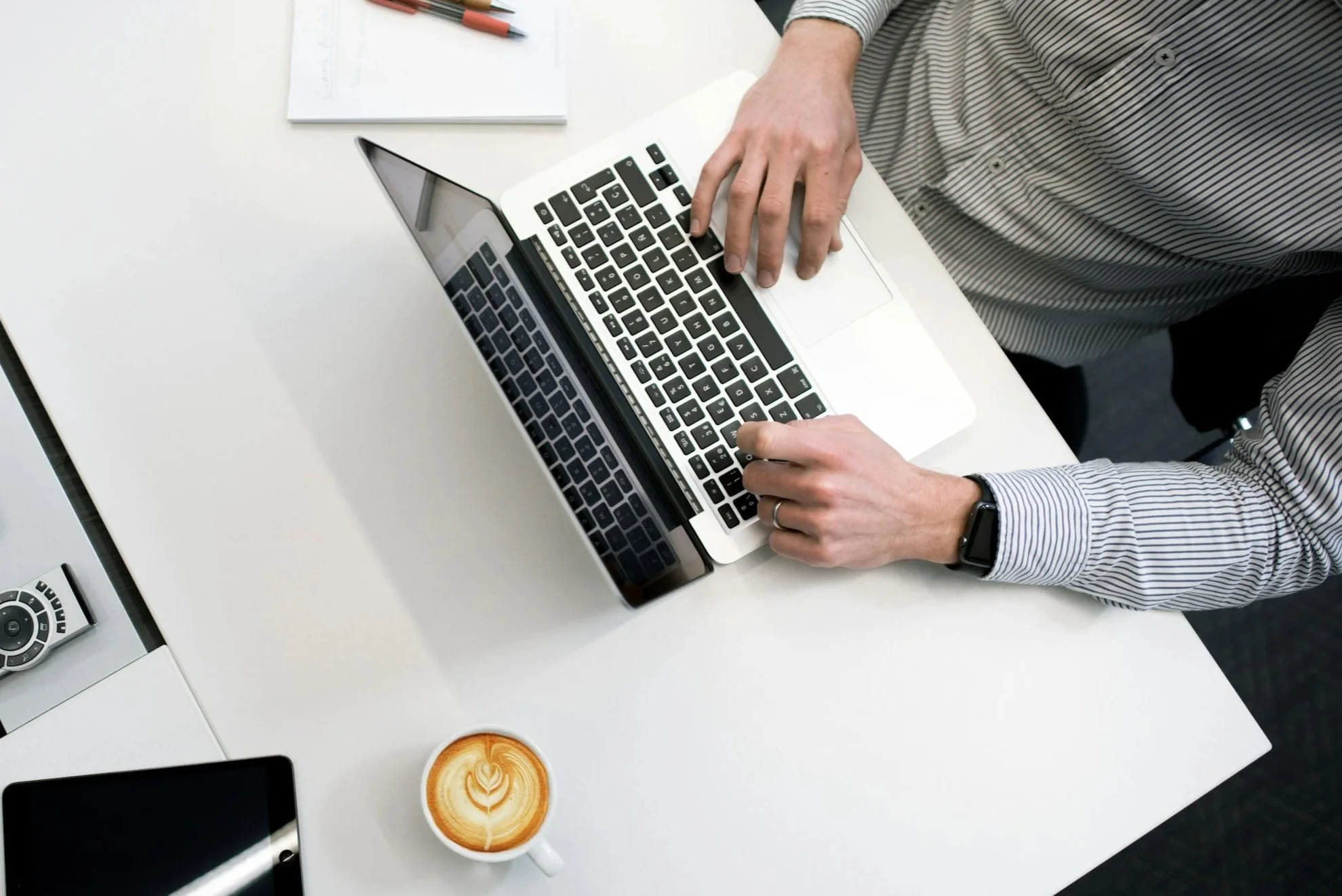 Person arbeitet an einem Laptop an einem weißen Schreibtisch, mit einem Notizbuch, Stiften, einem Smartphone, einem Kaffee und einem DVD-Player auf dem Tisch