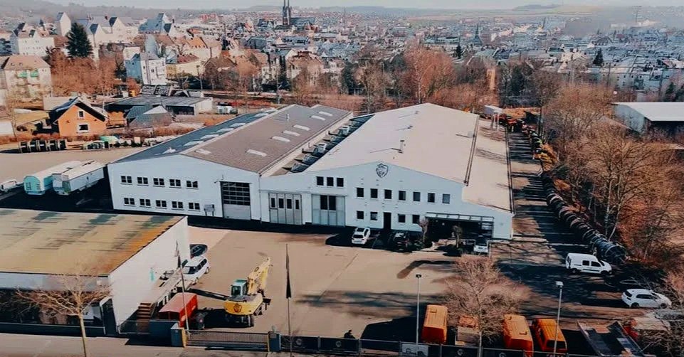 Luftaufnahme eines Industriegebäudes mit Parkplatz, umgeben von Bäumen und Wohnhausbebauung, bei Tageslicht.