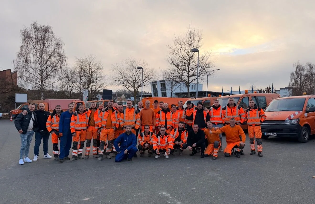 Ein Bild des Bee-d-Tech-Teams. Gruppe von Personen in orange Sicherheitskleidung vor mehreren orangefarbenen Fahrzeugen, draußen bei Sonnenuntergang.