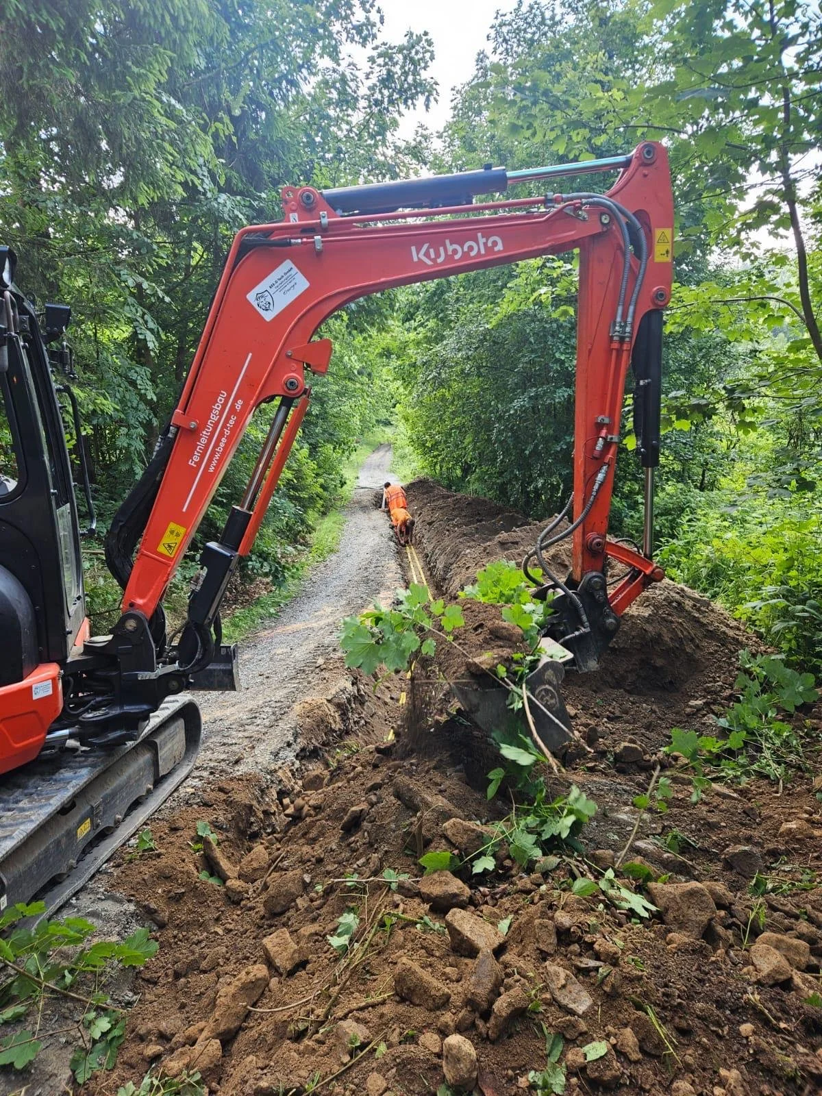 Ein Bagger arbeitet an einer Streckenbaustelle im Wald, hebt Erde und Steine aus einer Erdgrube, während zwei Arbeiter in orange Arbeitskleidung in der Erde graben.