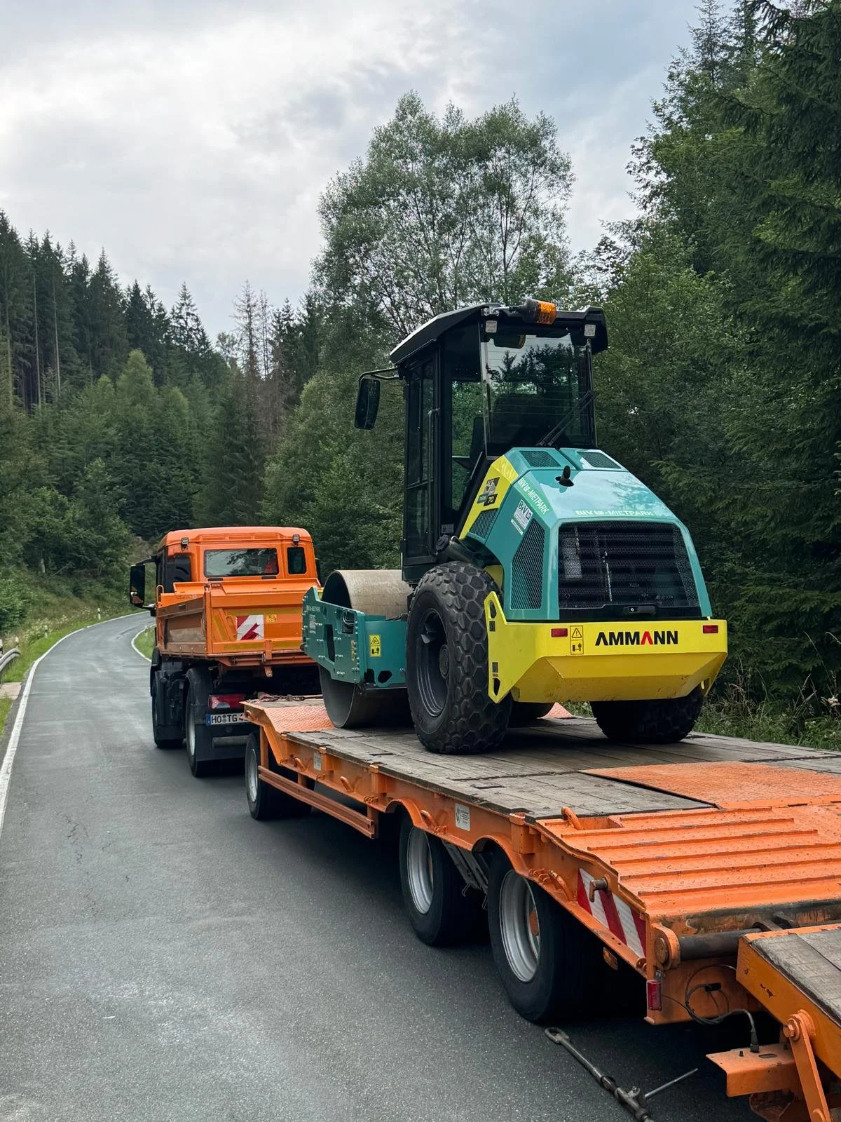 Ein Baugerät auf einem orangefarbenen Transportanhänger auf einer Landstraße, umgeben von Bäumen und Wald.