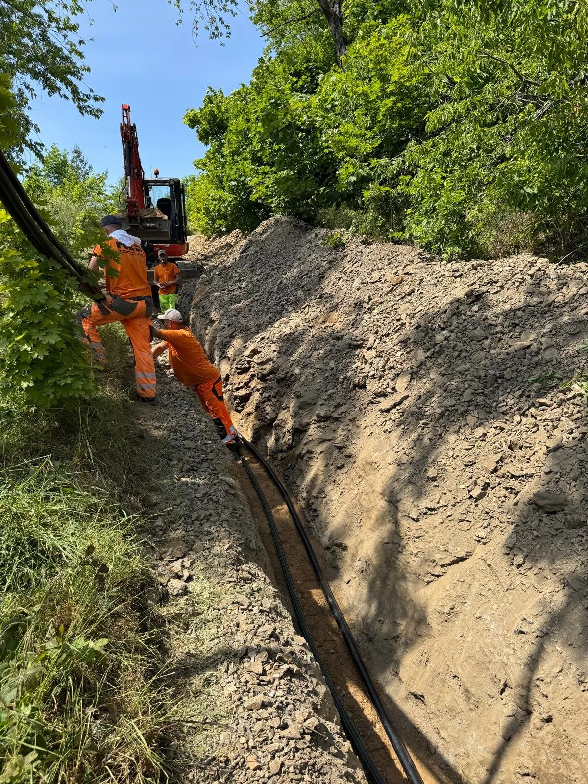 Arbeiten an einem Erdrutsch: Bauarbeiter legen Kabel in einem schlammigen Graben neben einer Baustelle mit Bagger, umgeben von grünen Bäumen.