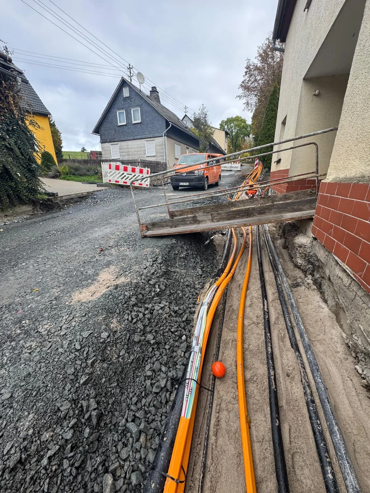 Straße mit Bauarbeiten, Kabel und Rohre verlaufen im Boden, Absperrung und Bauarbeiten sind sichtbar, im Hintergrund Häuser und ein orangefarbener Van.
