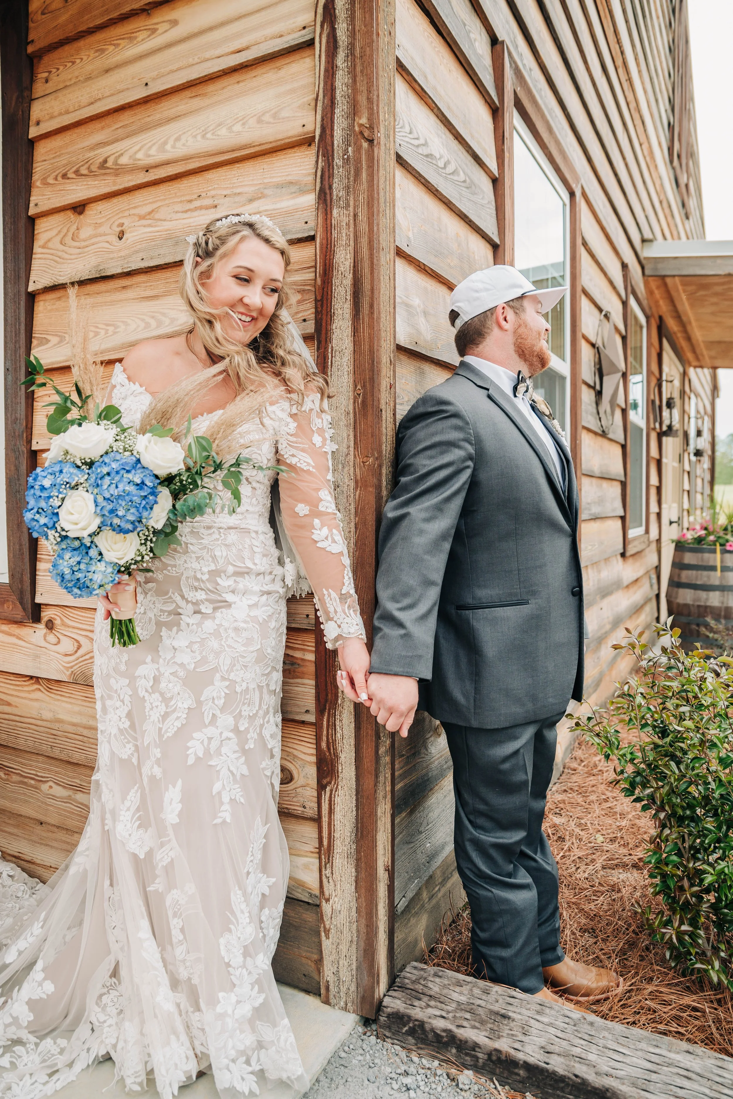Bride in a white lace wedding dress holding a bouquet of white and blue flowers, standing behind a groom in a gray suit and white cap, both holding hands and leaning against a wooden building.