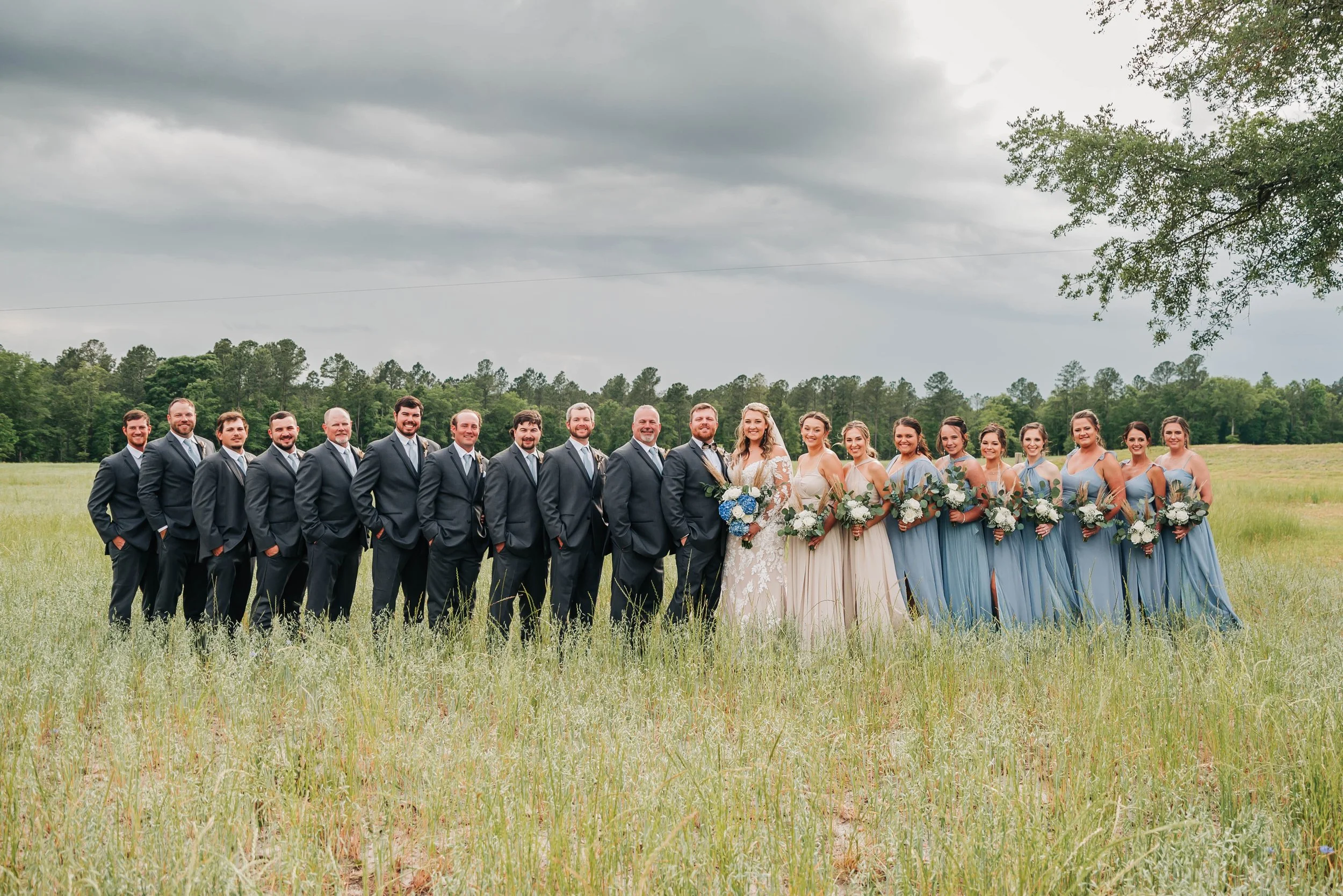 Wedding party standing outdoors in a grassy field on a cloudy day, with groomsmen on the left, bride and bridesmaids on the right, holding bouquets.