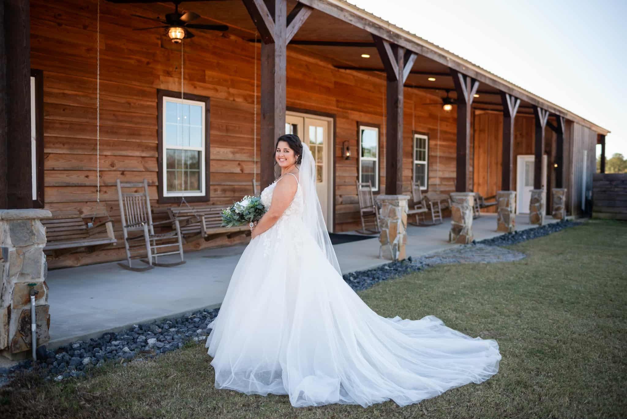 A bride in a white wedding gown holding a bouquet stands outside a rustic wooden building with rocking chairs on the porch.