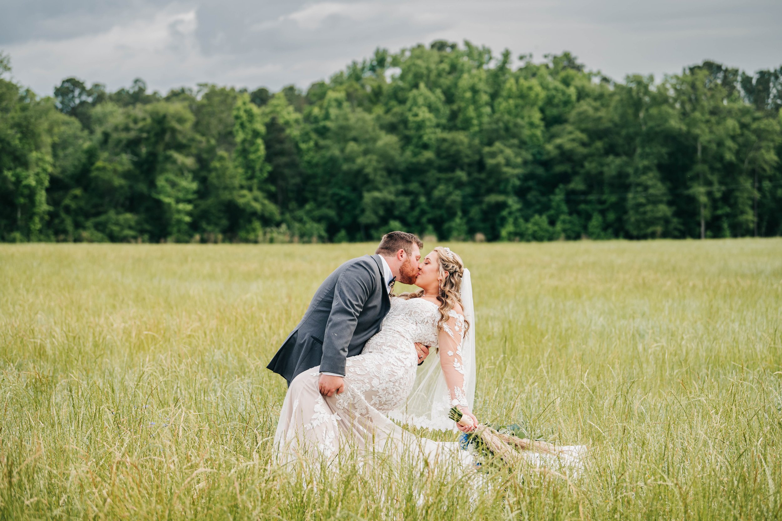 A bride and groom kiss in a grassy field; the groom leans forward to kiss the bride, who is holding a bouquet, with a background of green trees and a cloudy sky.