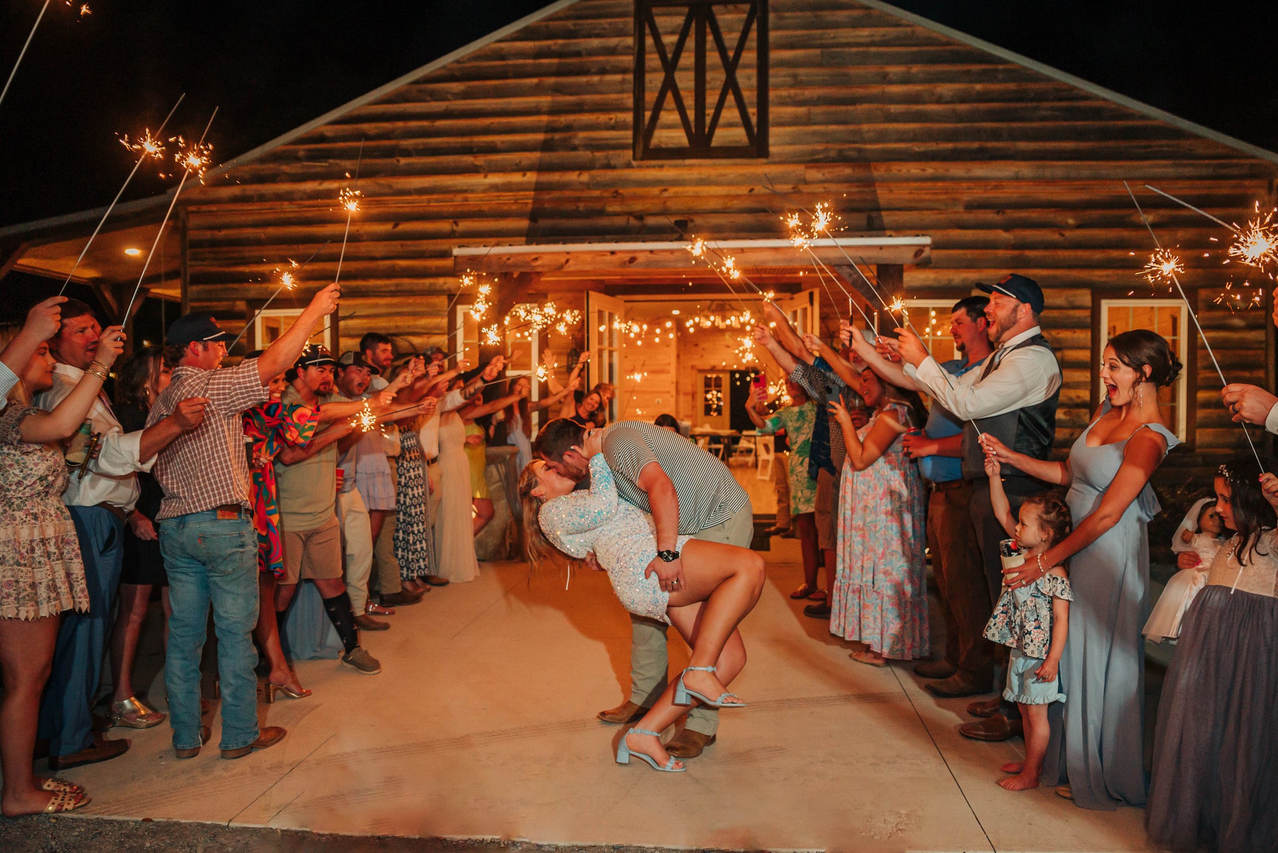 Couple dancing in the center while guests hold sparklers in a semi-circle behind them at a wedding celebration outside a rustic barn at night.