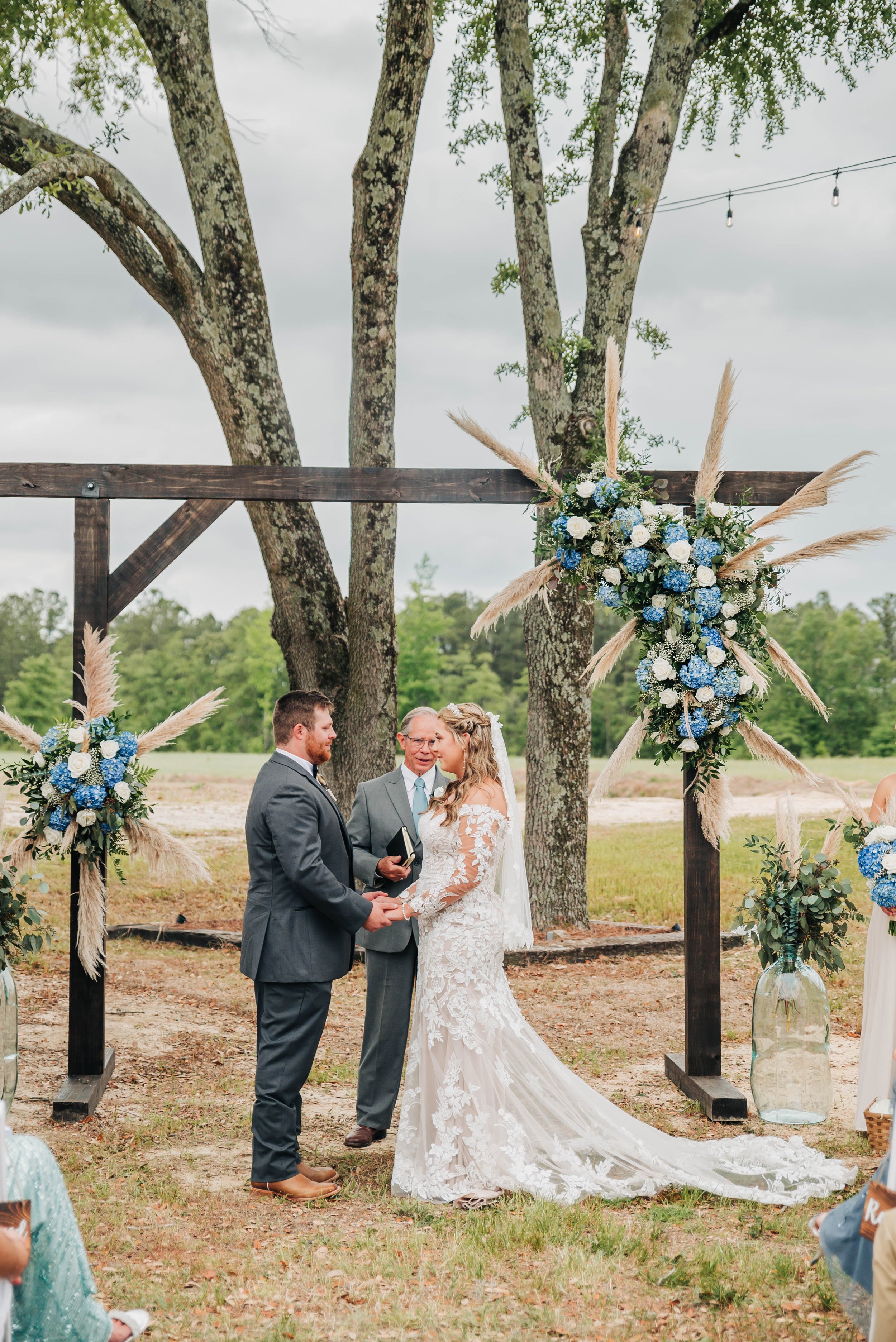 A couple getting married outdoors under a decorated arch, with an officiant in the middle, surrounded by trees and cloudy sky.