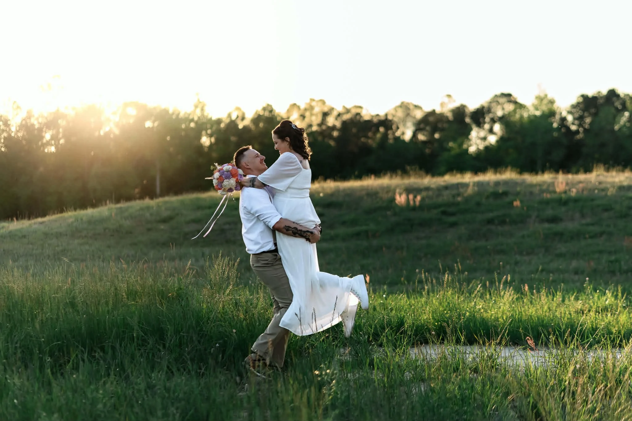 A couple, celebrating their wedding outdoors, with the groom holding the bride in his arms as they smile at each other during sunset in a green field.