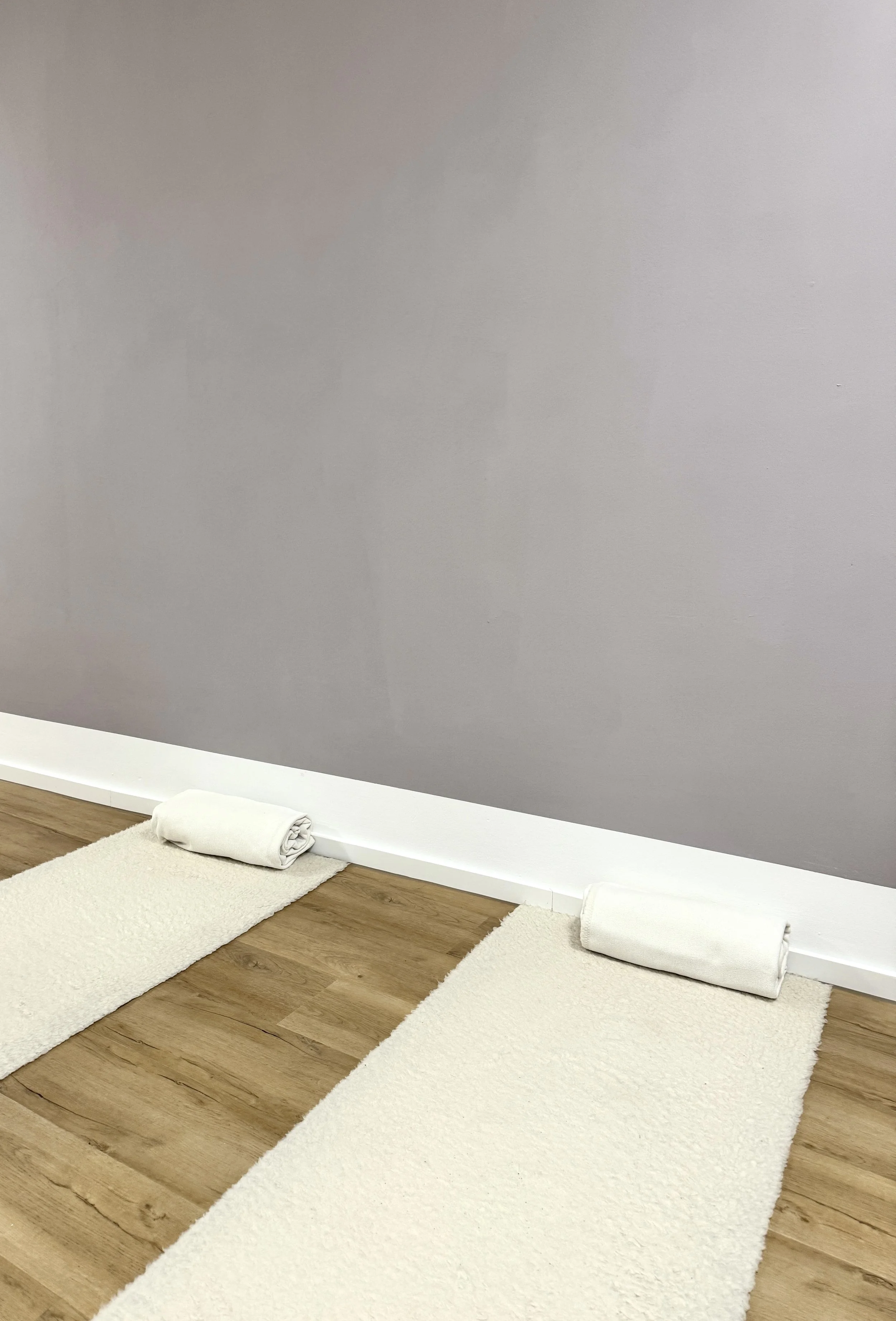 Zwei Yoga-Matten auf dem Boden neben einer grauen Wand, jeder mit einem weißen gefalteten Handtuch darauf.