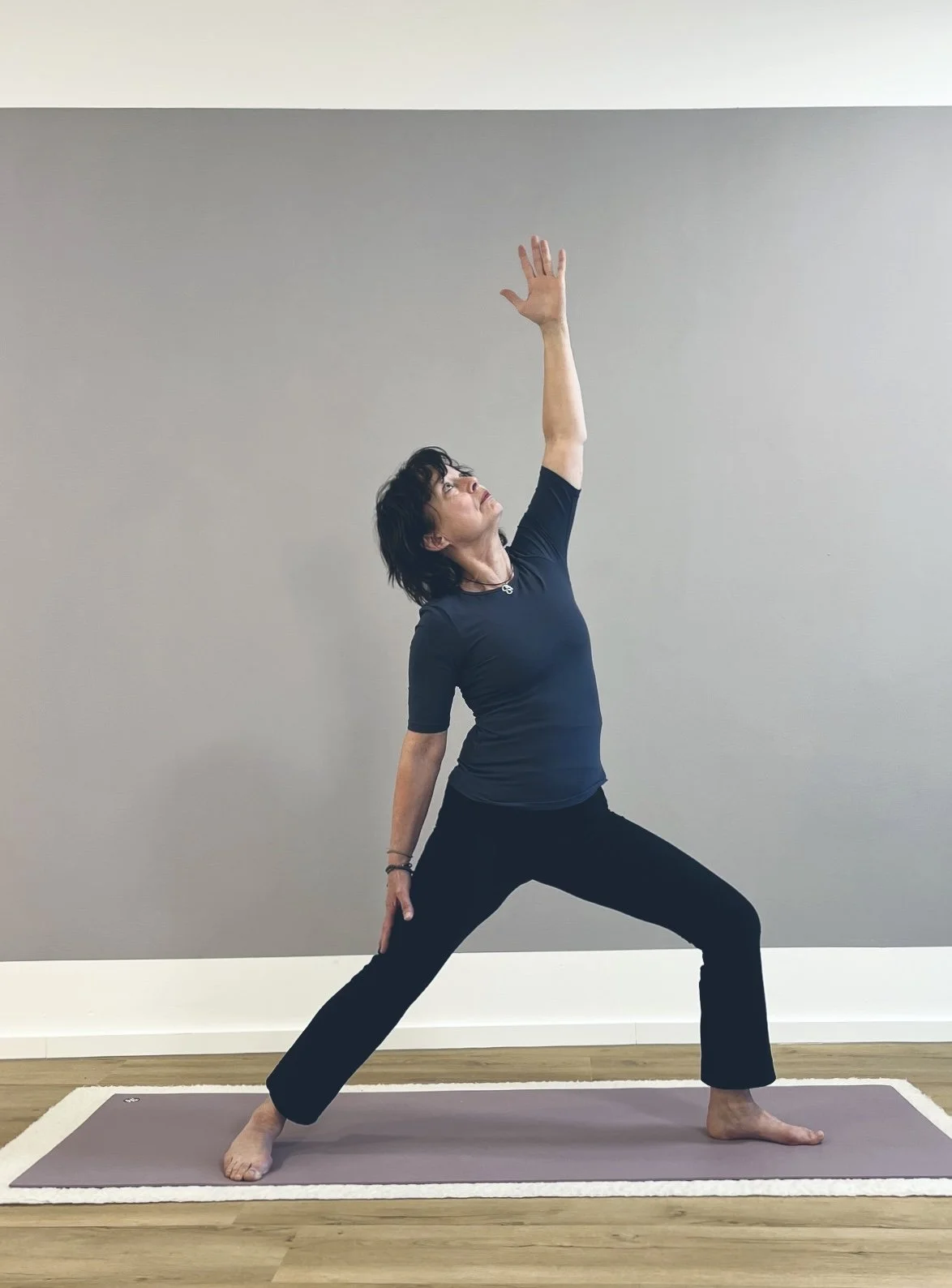 Frau in Yoga-Pose auf Yogamatte vor einer zweifarbigen Wand, hebt einen Arm und blickt nach oben.