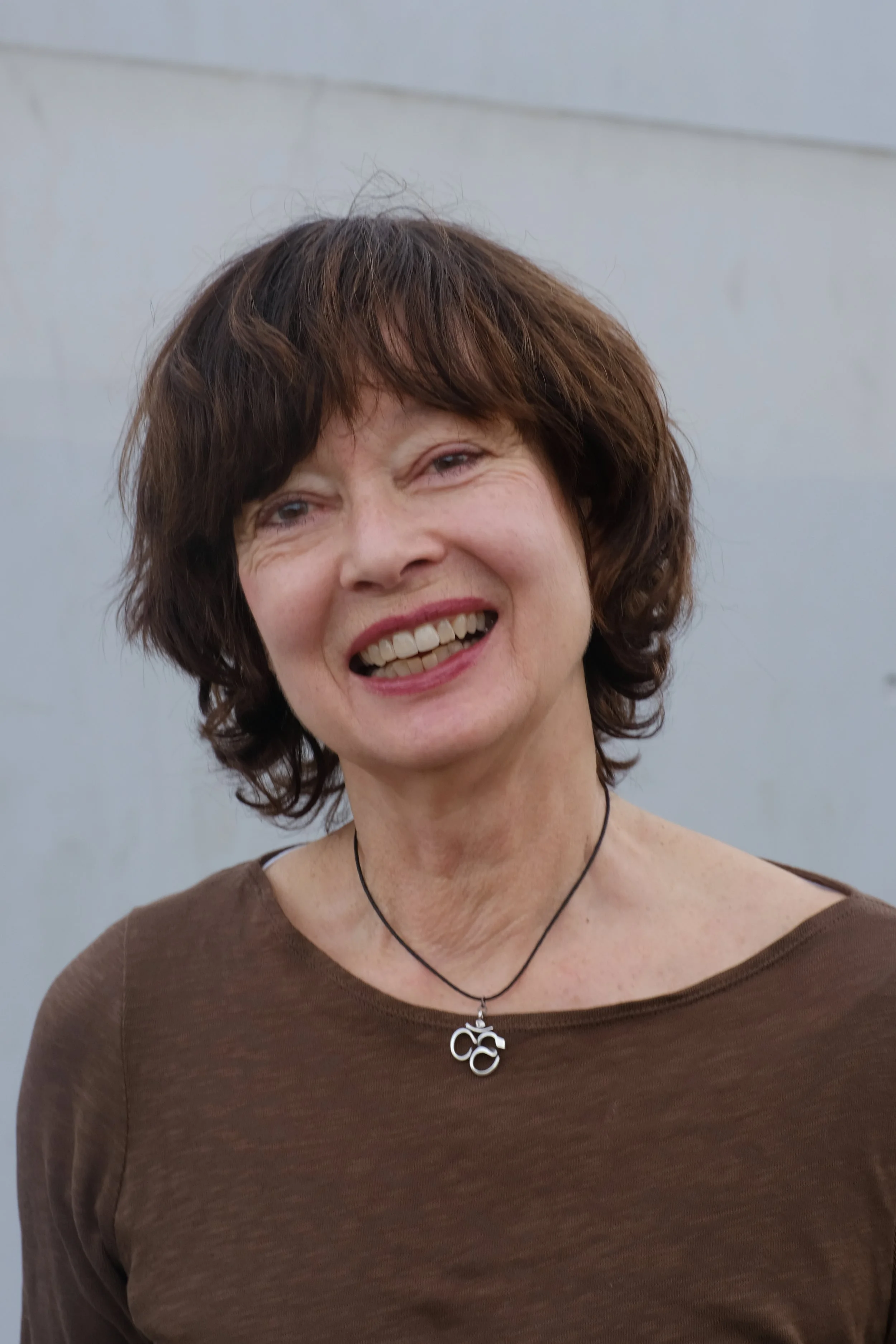 Eine lächelnde ältere Frau mit kurzen braunen Haaren, trägt ein braunes Oberteil und eine schwarze Halskette mit einem Anhänger, vor einem grauen Hintergrund.