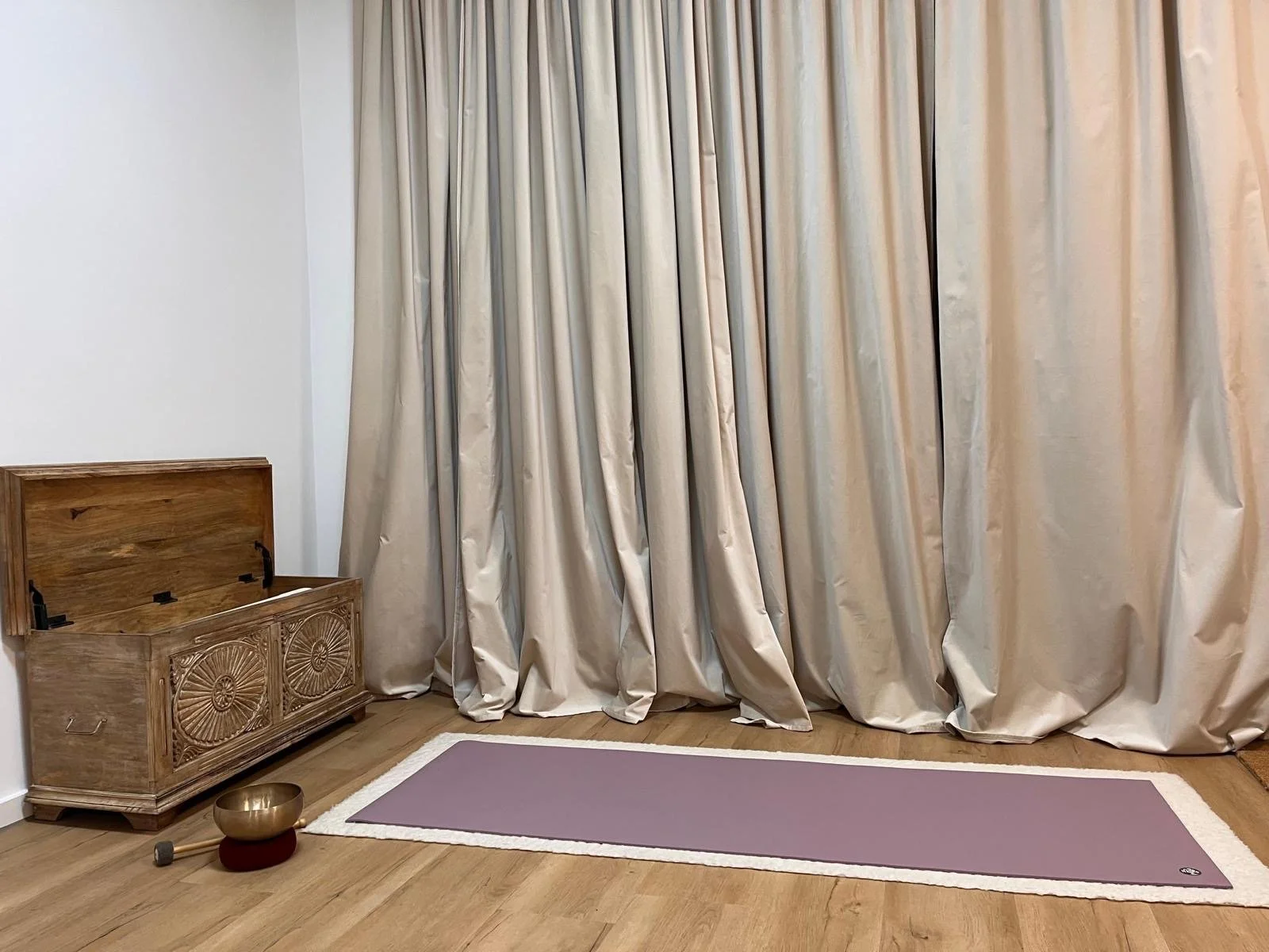 Ein Yoga-Raum mit blauer Matte auf Holzboden, großen beige Gardinen, einem geschnitzten Holztruhe in der Ecke, einem glänzenden tibetischen Klangschalen-Set auf dem Boden.