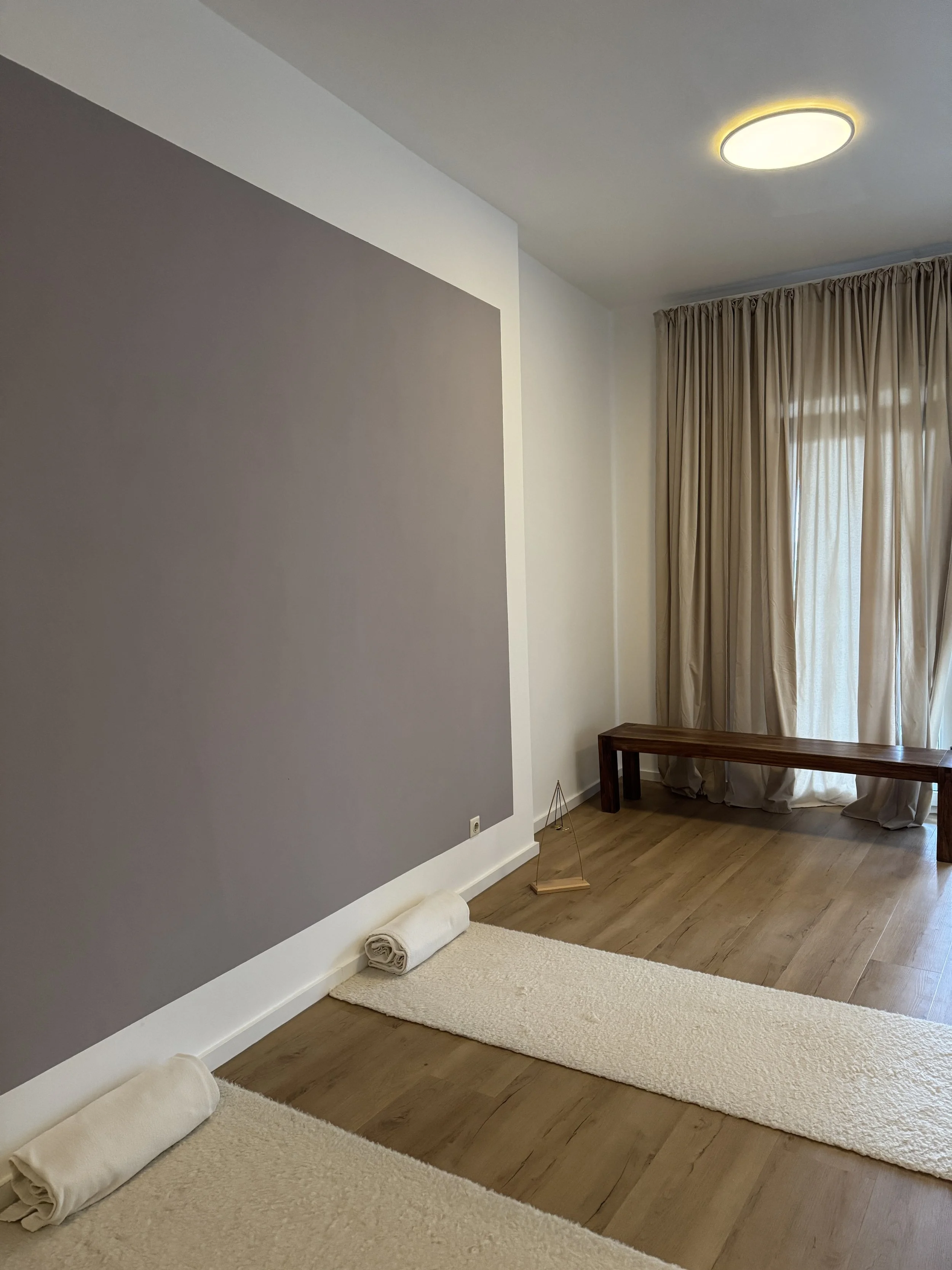 Ein ruhiger, minimalistisch eingerichteter Raum mit Holzfußboden, langen cremefarbenen Vorhängen, samtfarbenen Läufern auf dem Boden, einem braunen Holzbank und einer Wand mit grauer Akzentwand.