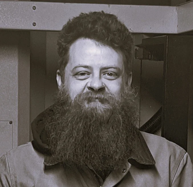 Black-and-white photo of a man with a full beard and moustache, smiling, wearing a Carhartt jacket, in front of a CNC machine.