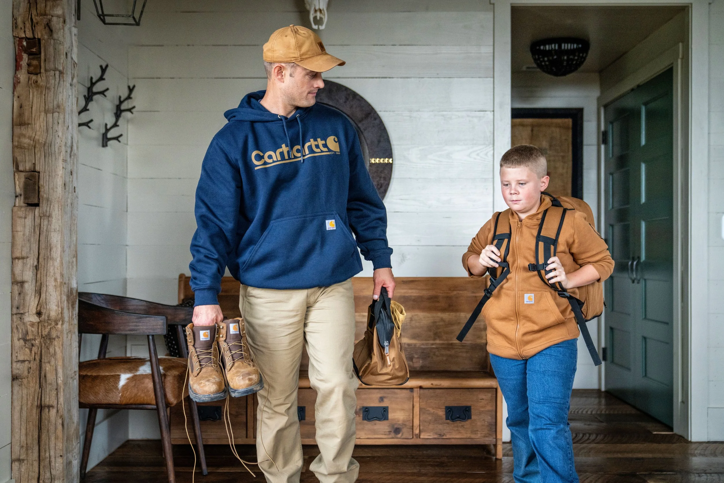 Father and his son leaving the house wearing Carhartt sweatshirts, carrying Carhartt boots and a backpack.