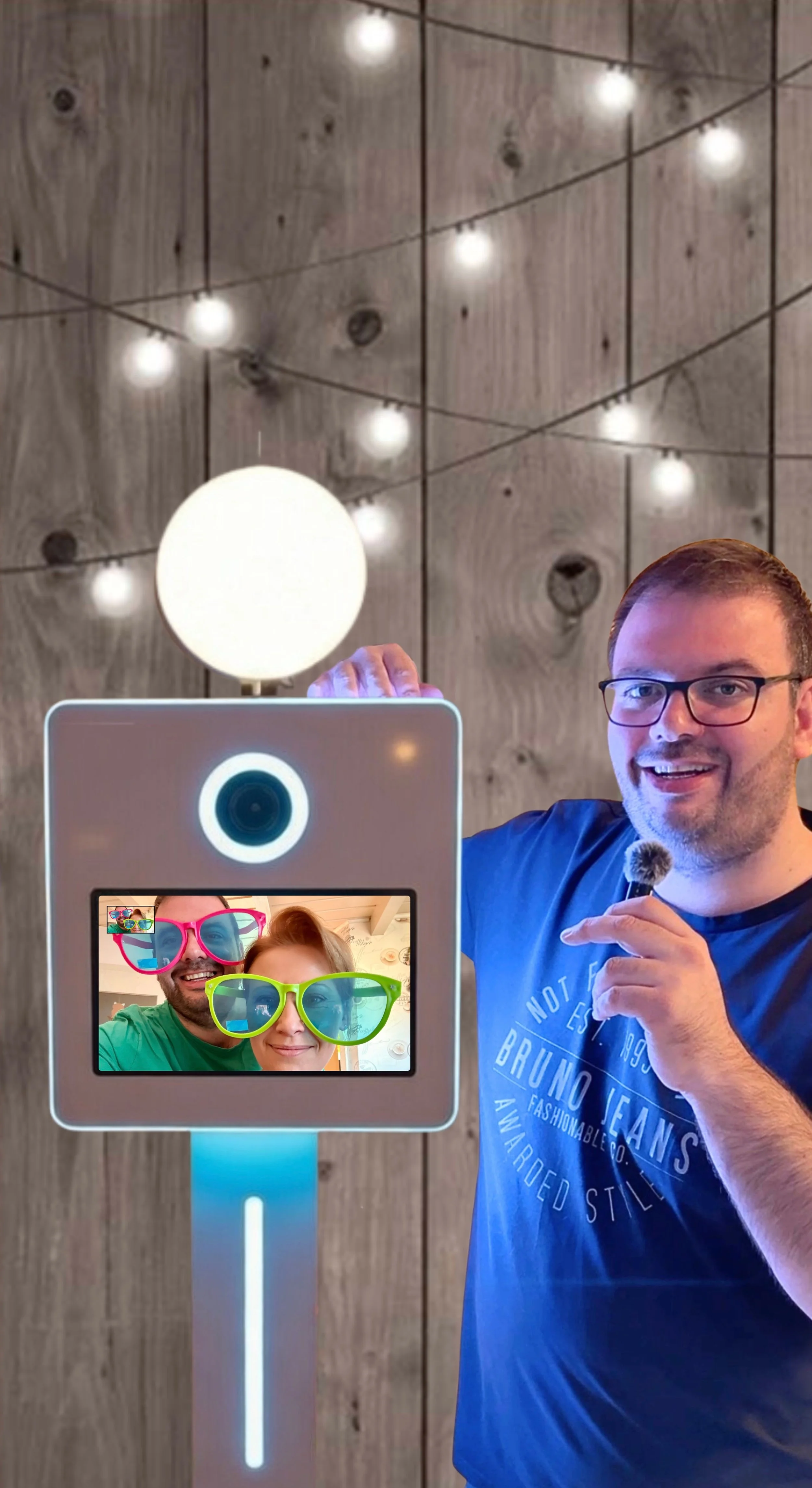 Ein Mann macht ein Selfie mit einer Fotobox. Im Hintergrund sind Lichter an einer Holzwand sichtbar.