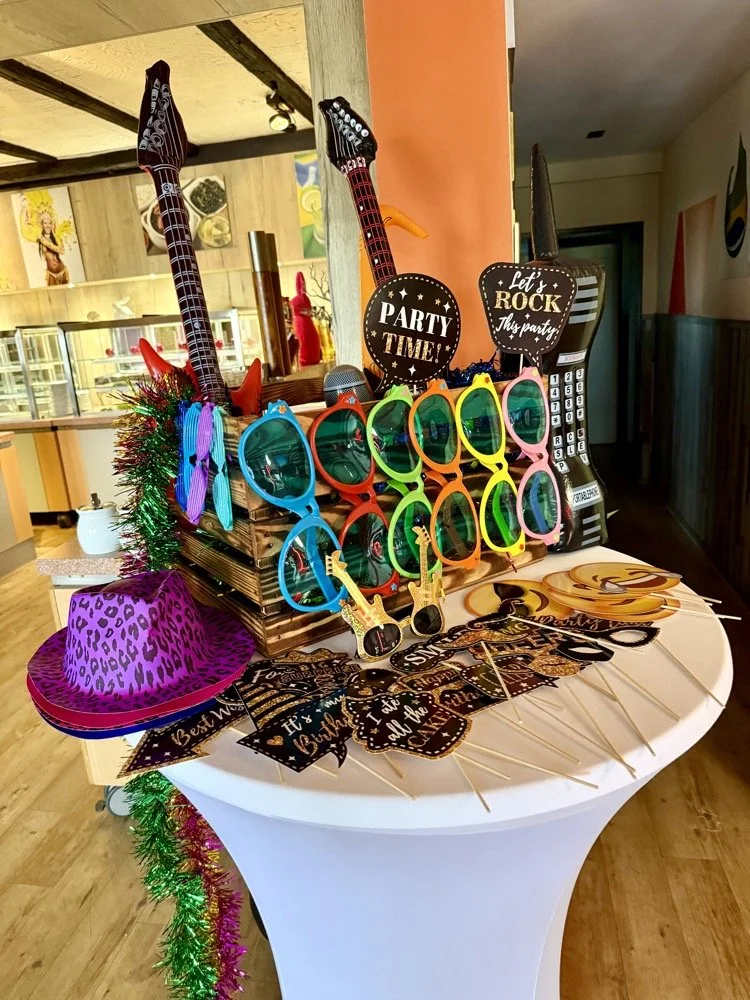 Dekorative Partyartikel auf einem runden Tisch, darunter bunte Sonnenbrillen, Mini-Gitarren, Partyhüte, und Schilder mit Party-Sprüchen wie "Party Time!" und "Let’s Rock this party". Im Hintergrund eine Bar mit Getränken und ein Spiegel.