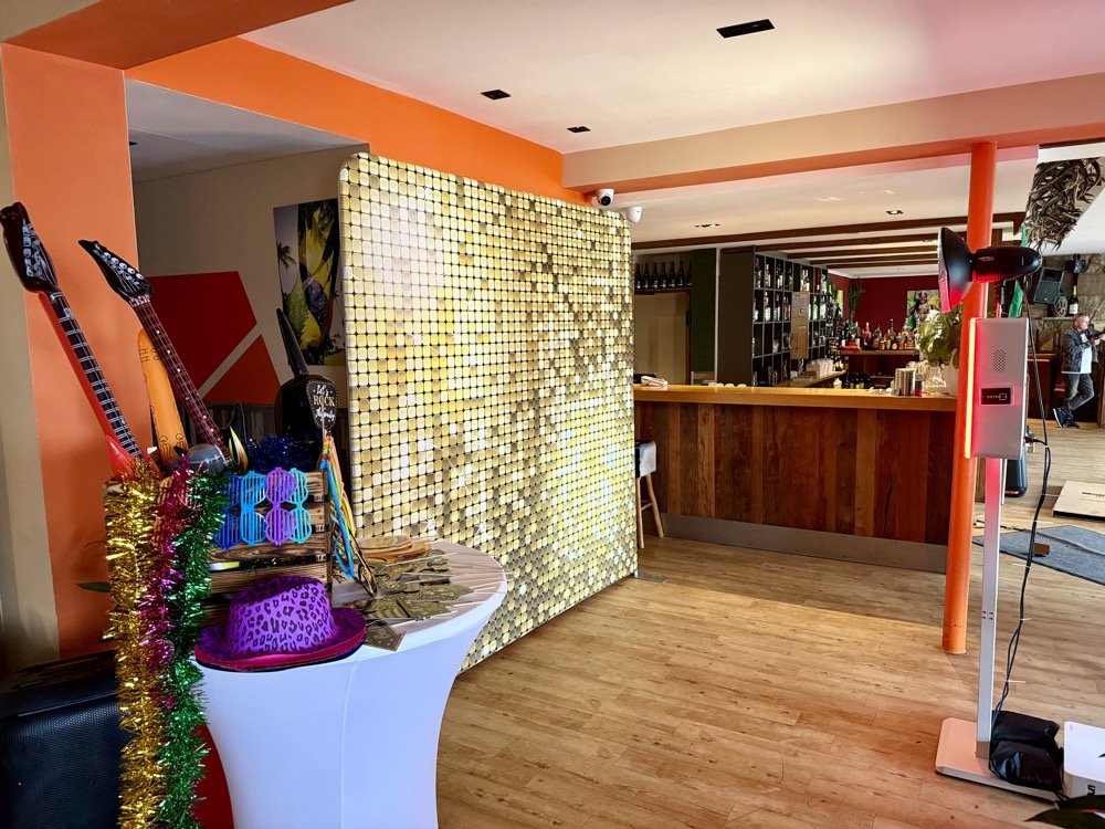 Innenaufnahme einer Bar mit Holztheke, dekoriert für eine Feier, mit Glitzertapete, Musik- und Partyzubehör, und einem orangefarbenen Wandabschnitt.