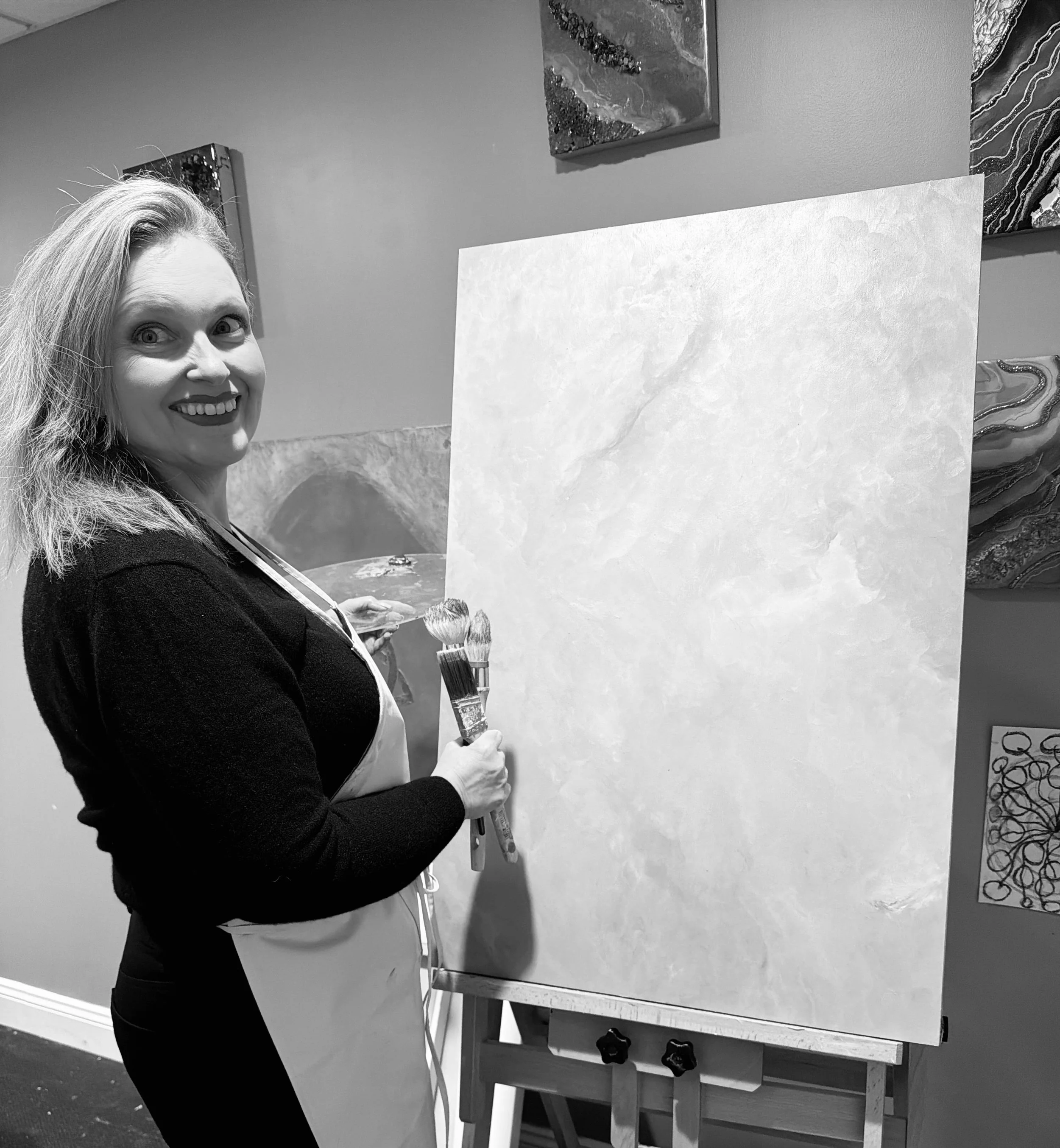 Tatyana Y. Kravchenko is smiling while holding paintbrushes in front of a canvas in an art studio.