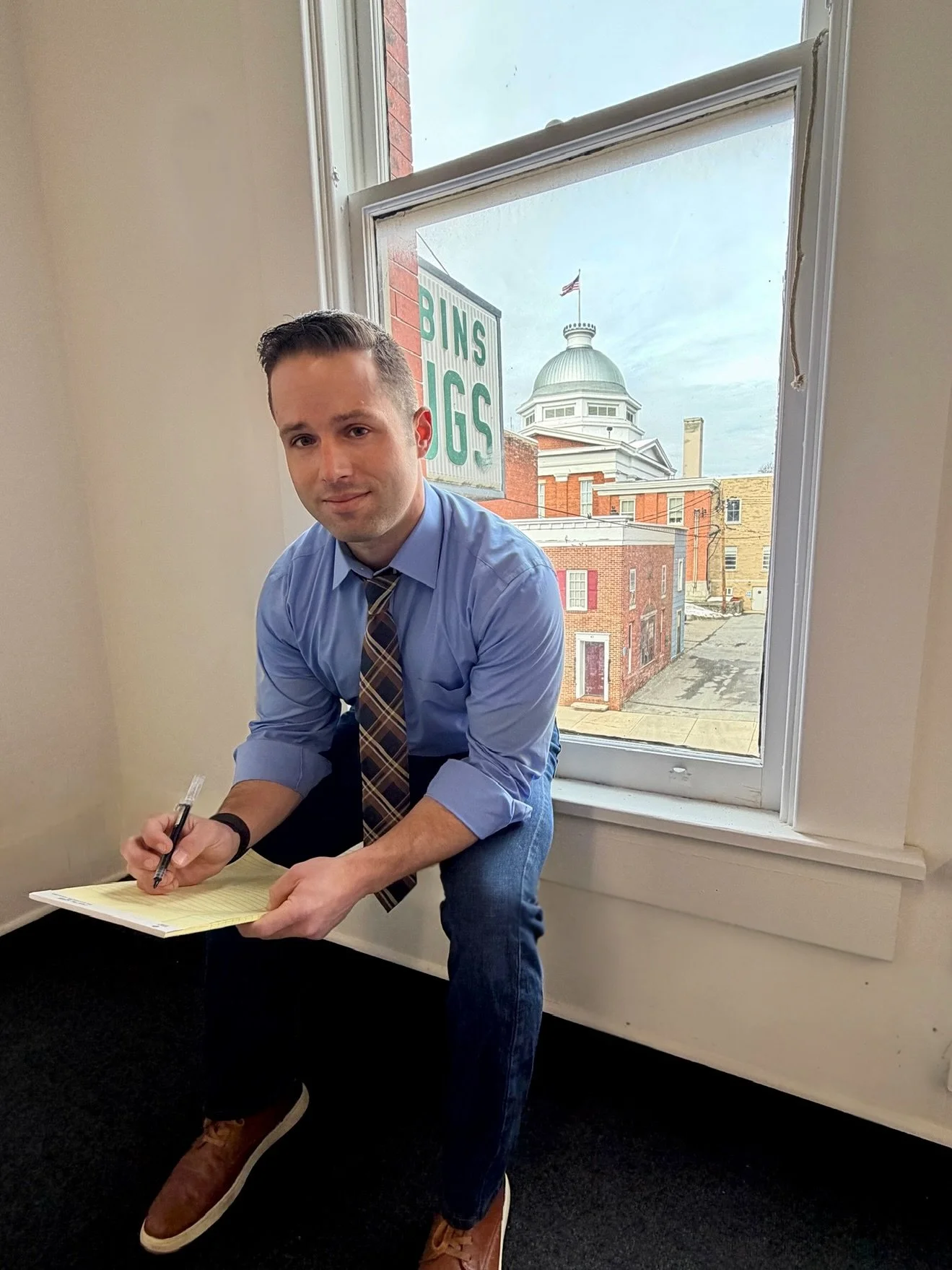 A man in a blue shirt and plaid tie squatting near a window, holding a yellow legal pad and pen, with a cityscape view including a courthouse and a sign that reads 'BINS JUGS' behind him.