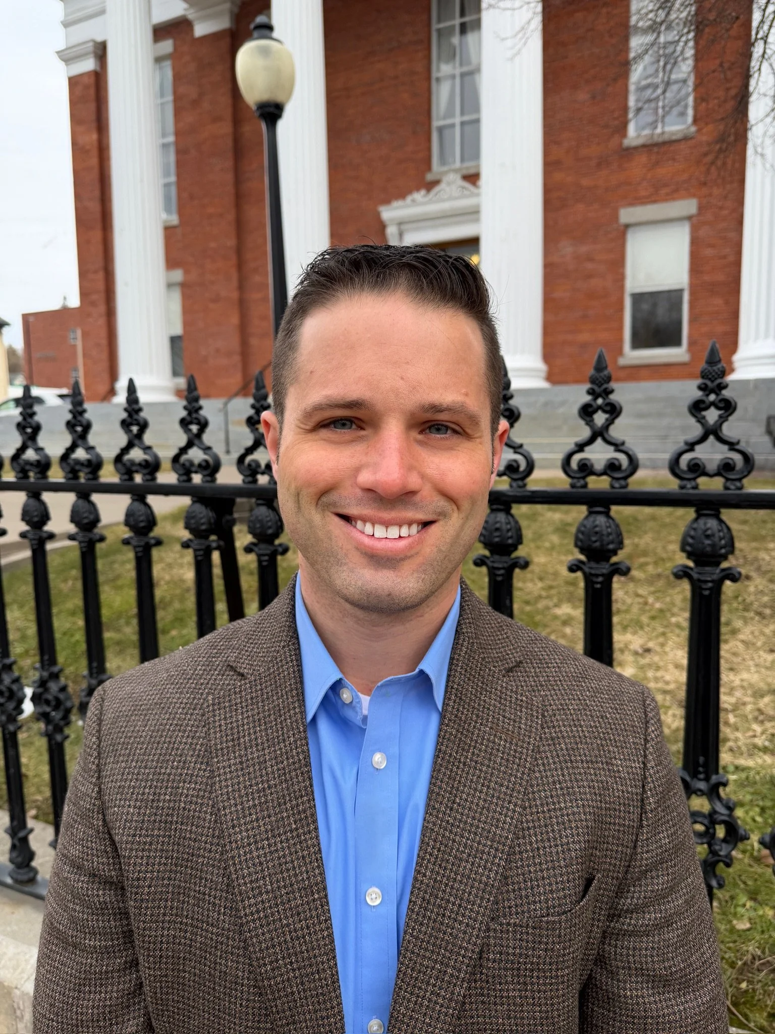 A smiling man in a blue shirt and brown blazer standing outside in front of a black iron fence and a brick building with white columns.