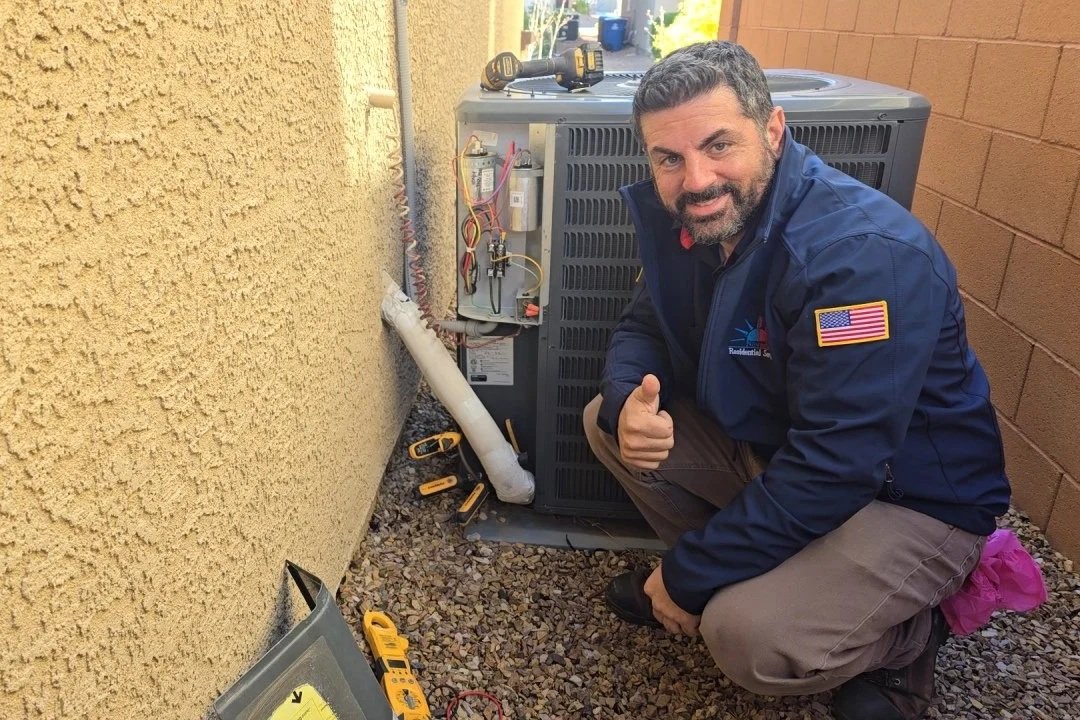 A man with a patch of the American flag on his sleeve crouching next to an HVAC unit outdoors, giving a thumbs-up.