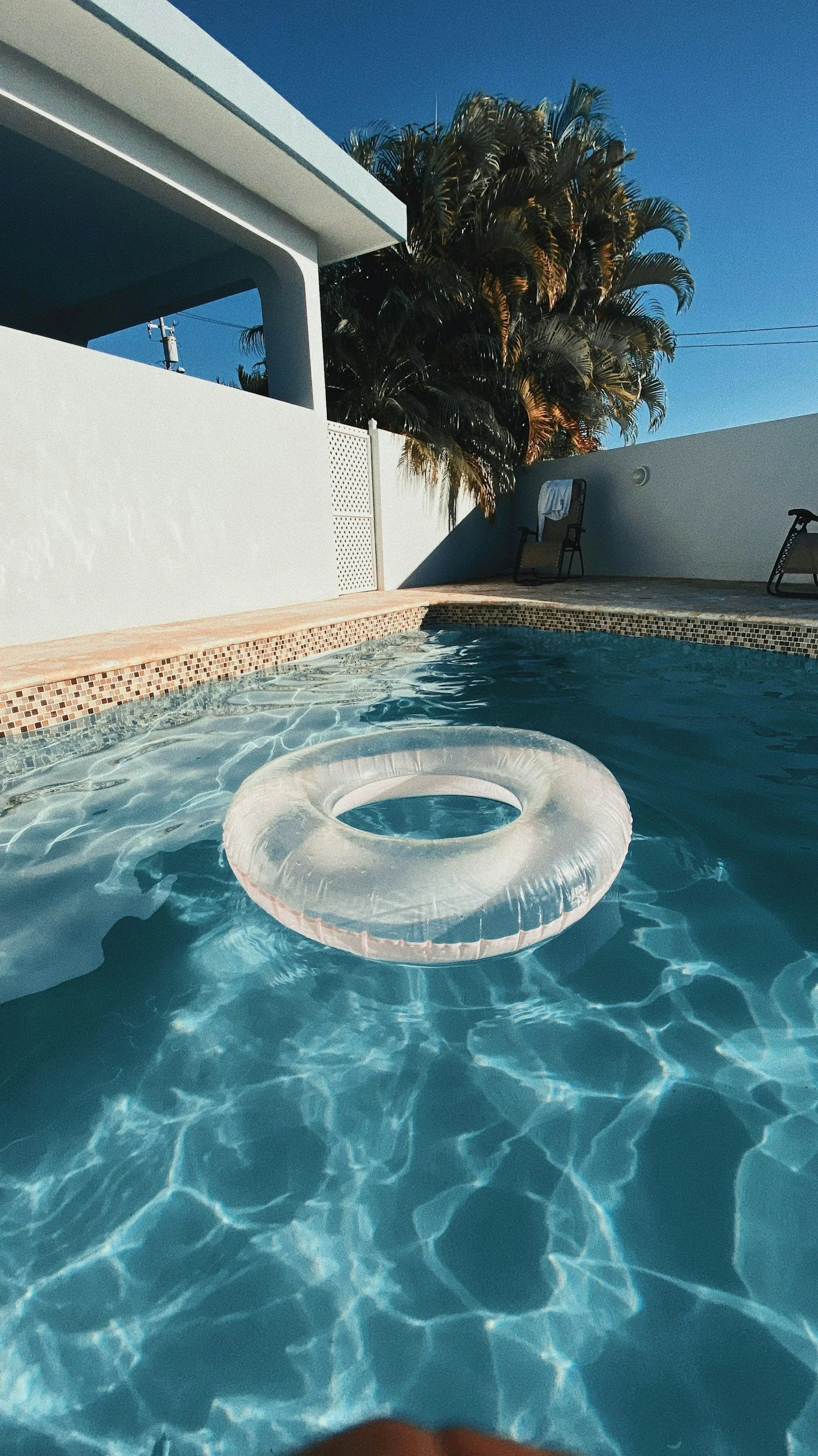 A swimming pool with a clear inflatable ring float in the water, surrounded by a white patio wall, with two lounge chairs and palm trees in the background under a clear blue sky.