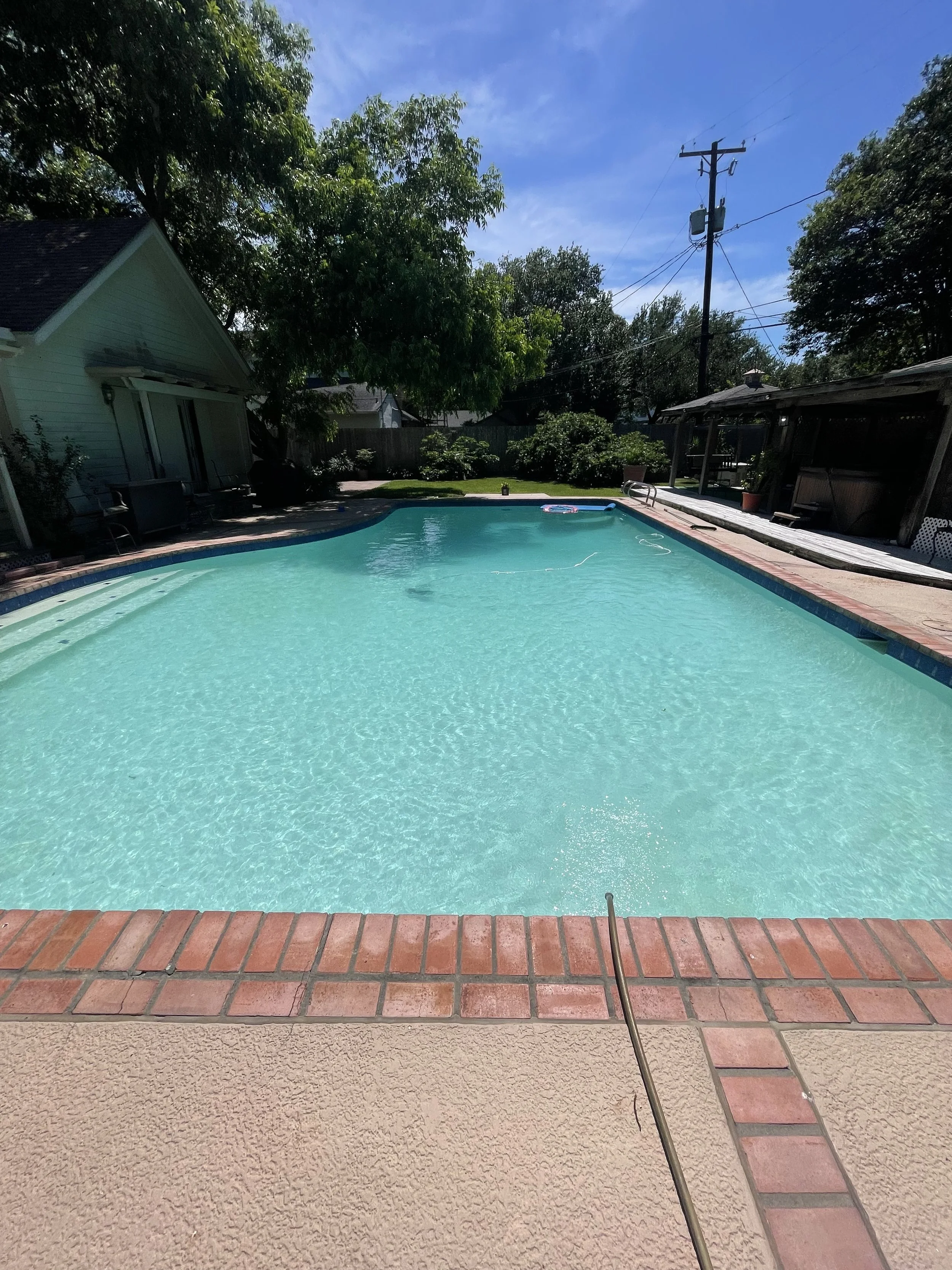 Backyard swimming pool with clear blue water, surrounded by a brick and concrete deck, trees, and a fence under a partly cloudy sky.