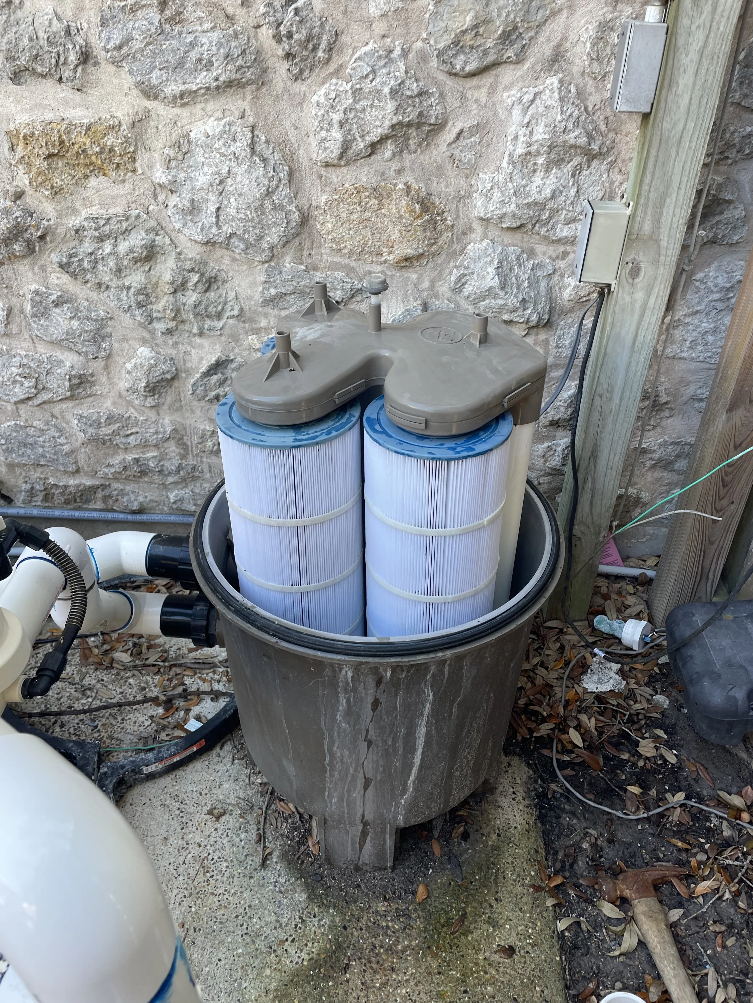 A pool filtration system with two large white filter cartridges installed inside a gray filter housing, set outside against a stone wall, with various pipes, wires, and tools around it.