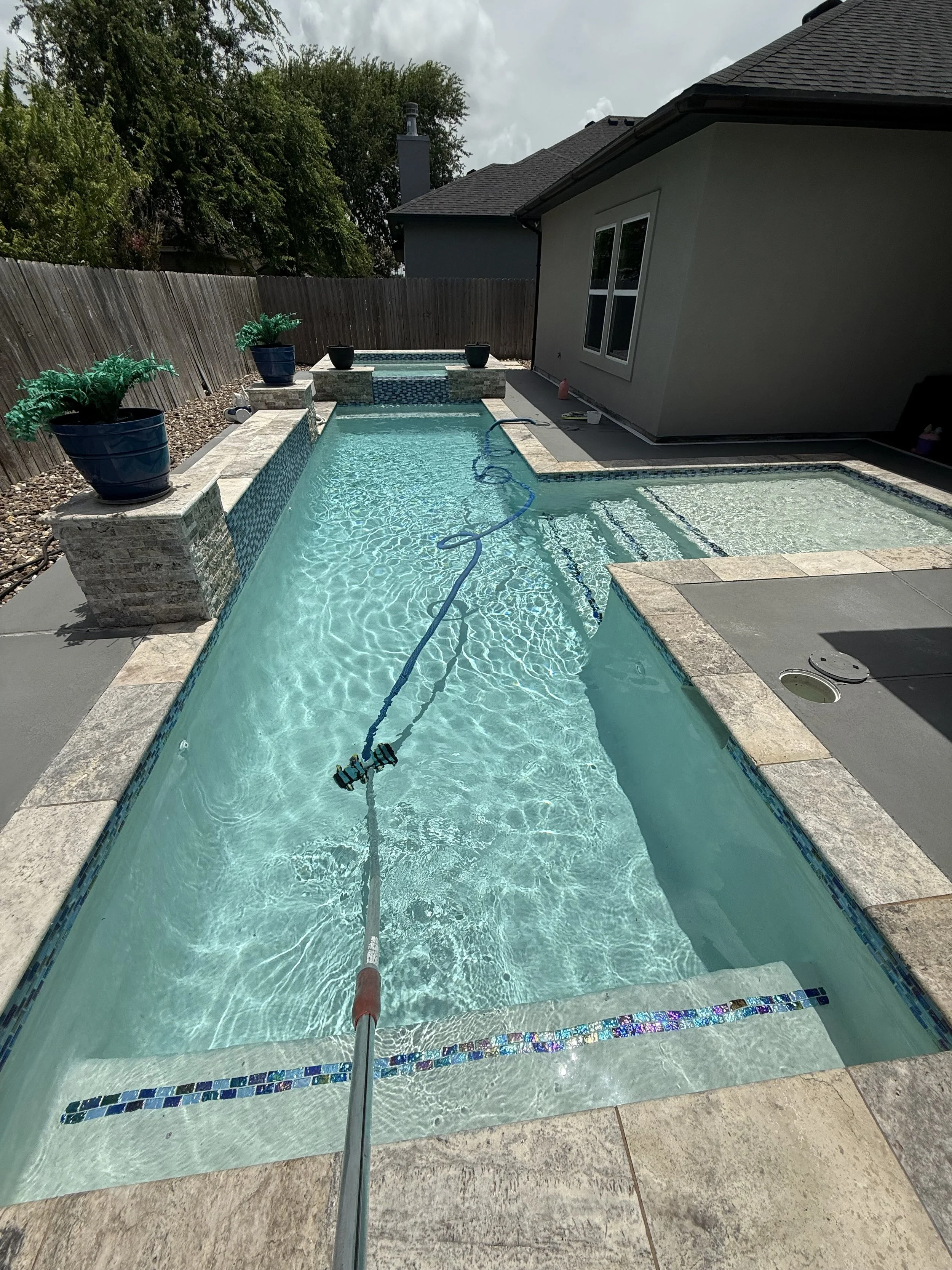 A backyard swimming pool with a cleaning pole extended into the water. The pool has tiled edges, a small attached hot tub, and is surrounded by potted plants, concrete, and a wooden fence. There is a house with a window nearby.