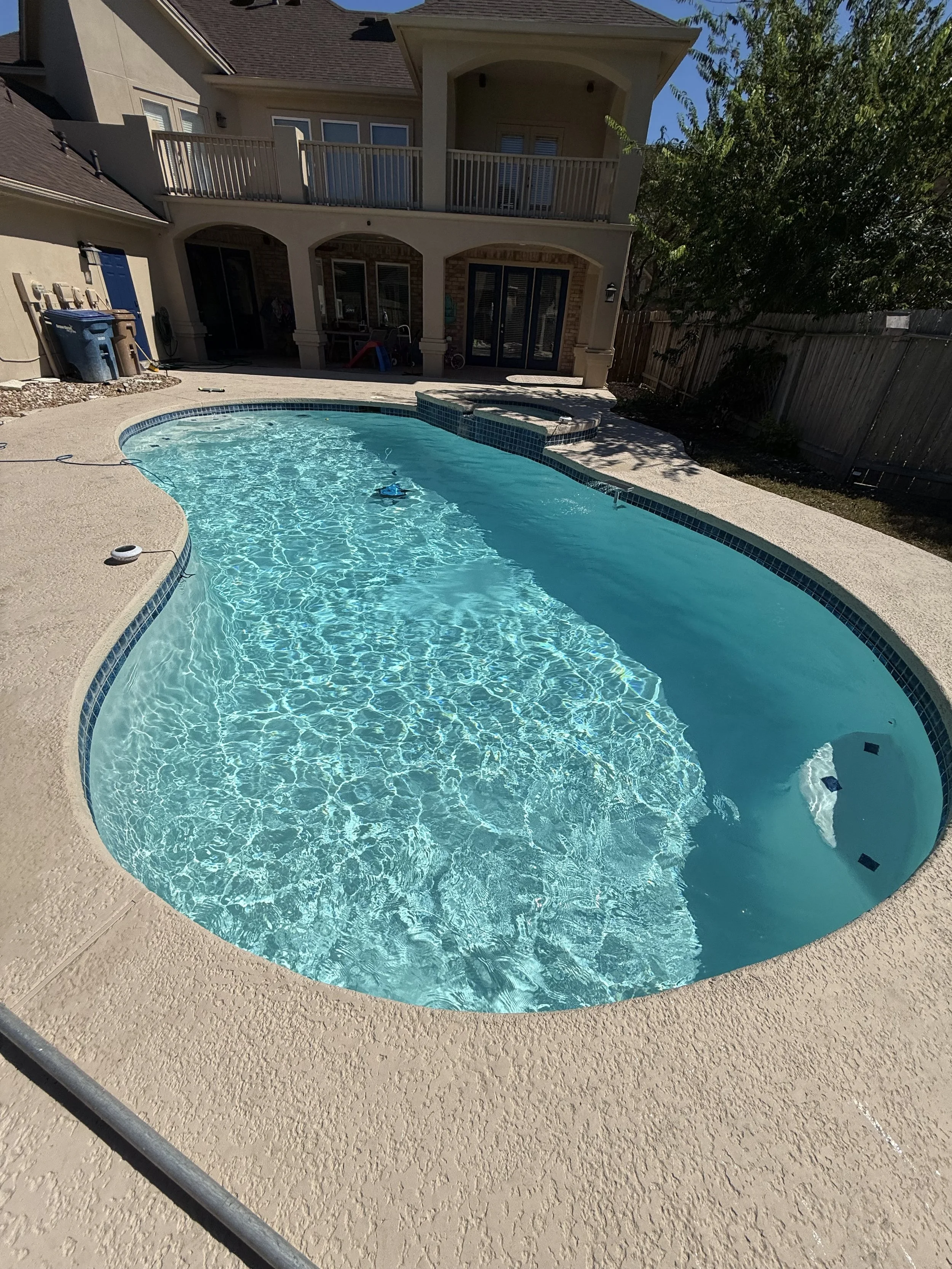 A backyard swimming pool with clear blue water, a hot tub, and a concrete deck, with a two-story house in the background, a wooden fence, and some trees on the right side.