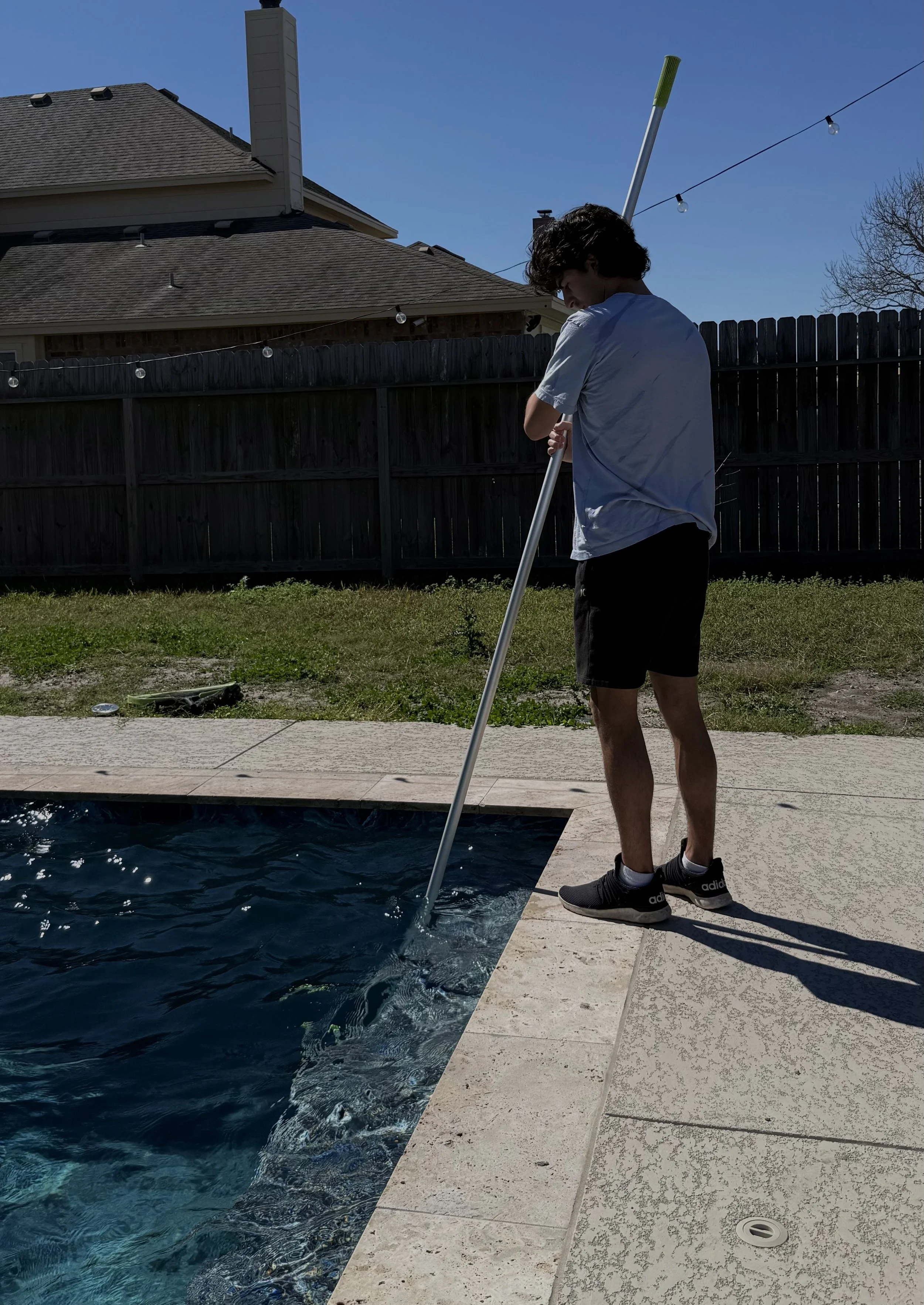 A person standing on the pool's edge holding a pool net, looking into the water on a sunny day.