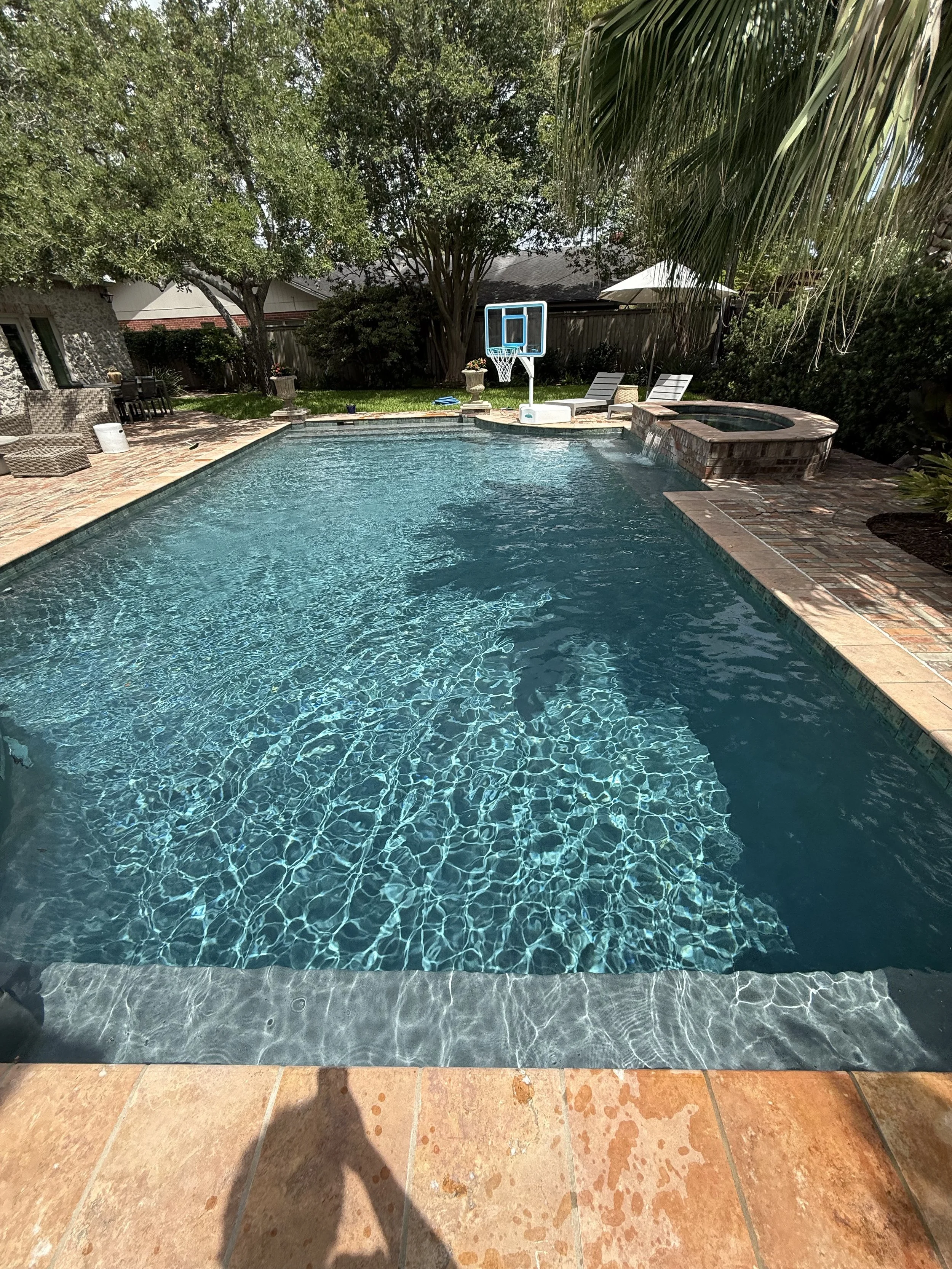 A backyard swimming pool with clear blue water, surrounded by a brick patio. There is a basketball hoop at one end of the pool, and a hot tub adjacent to it. Two lounge chairs are on the patio, with trees and shrubs framing the area.