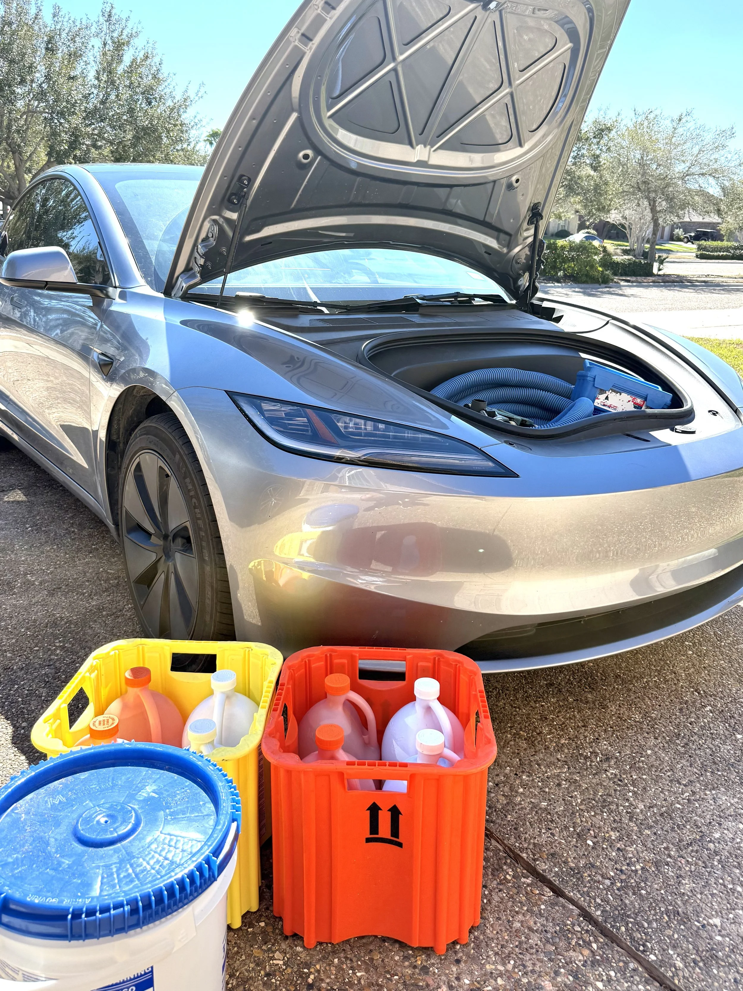 An electric car with its front hood open, revealing a blue hose inside. In front of the car, there are three large containers of cleaning or washing supplies, including a yellow basket with orange bottles, an orange basket with white bottles, and a blue and white bucket. The scene is outdoors on a driveway with trees and houses in the background.