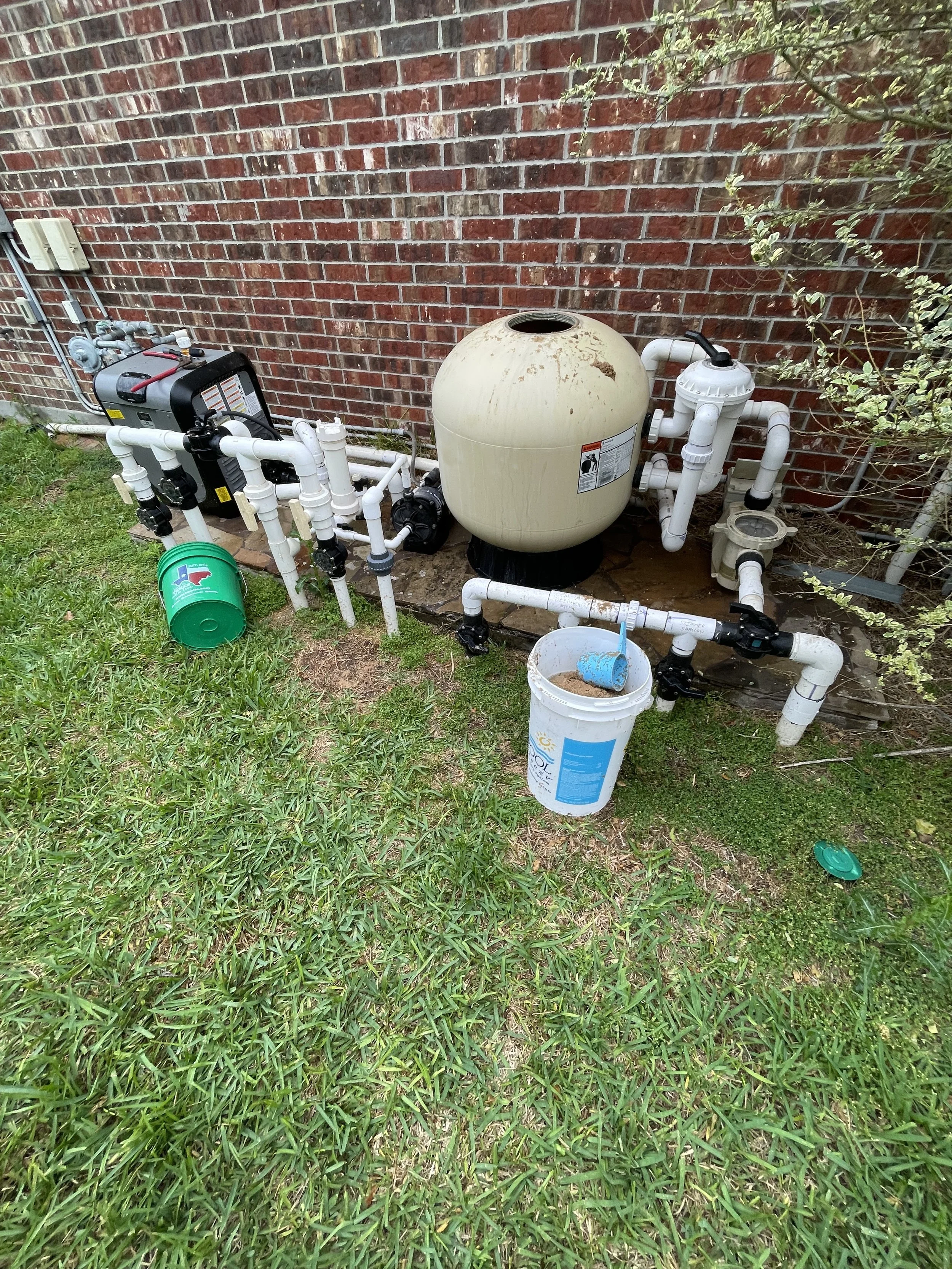 A backyard pool filter system with white PVC pipes, a large beige sand filter, a green bucket, and a bucket with sand and a blue tool, next to a red brick wall and green grass.