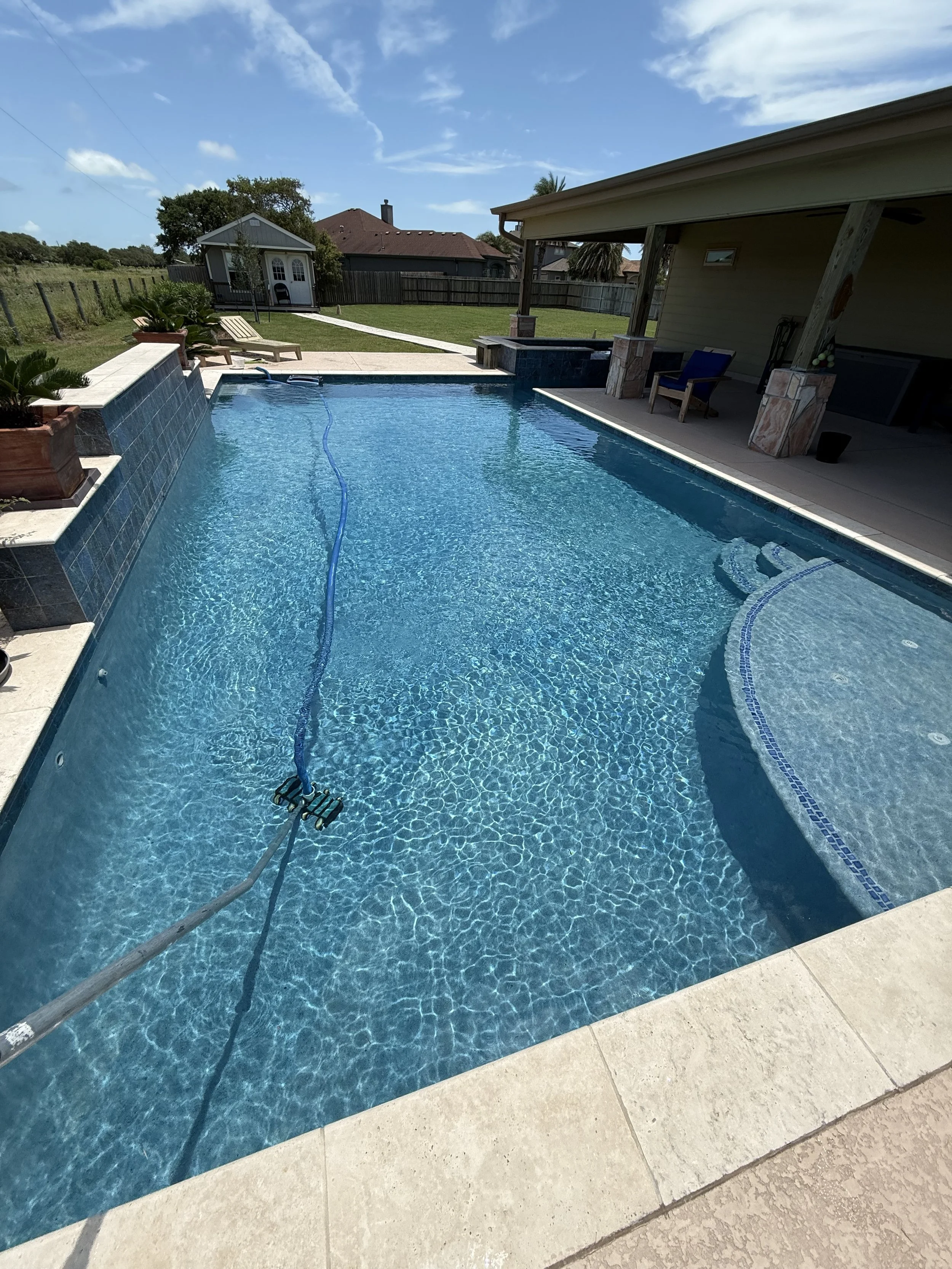 A backyard swimming pool with clear blue water, surrounded by a concrete deck, with pool cleaning equipment submerged in the water. In the background, there is a grassy area with a small building, lounge chairs, and a fence under a partly cloudy sky.