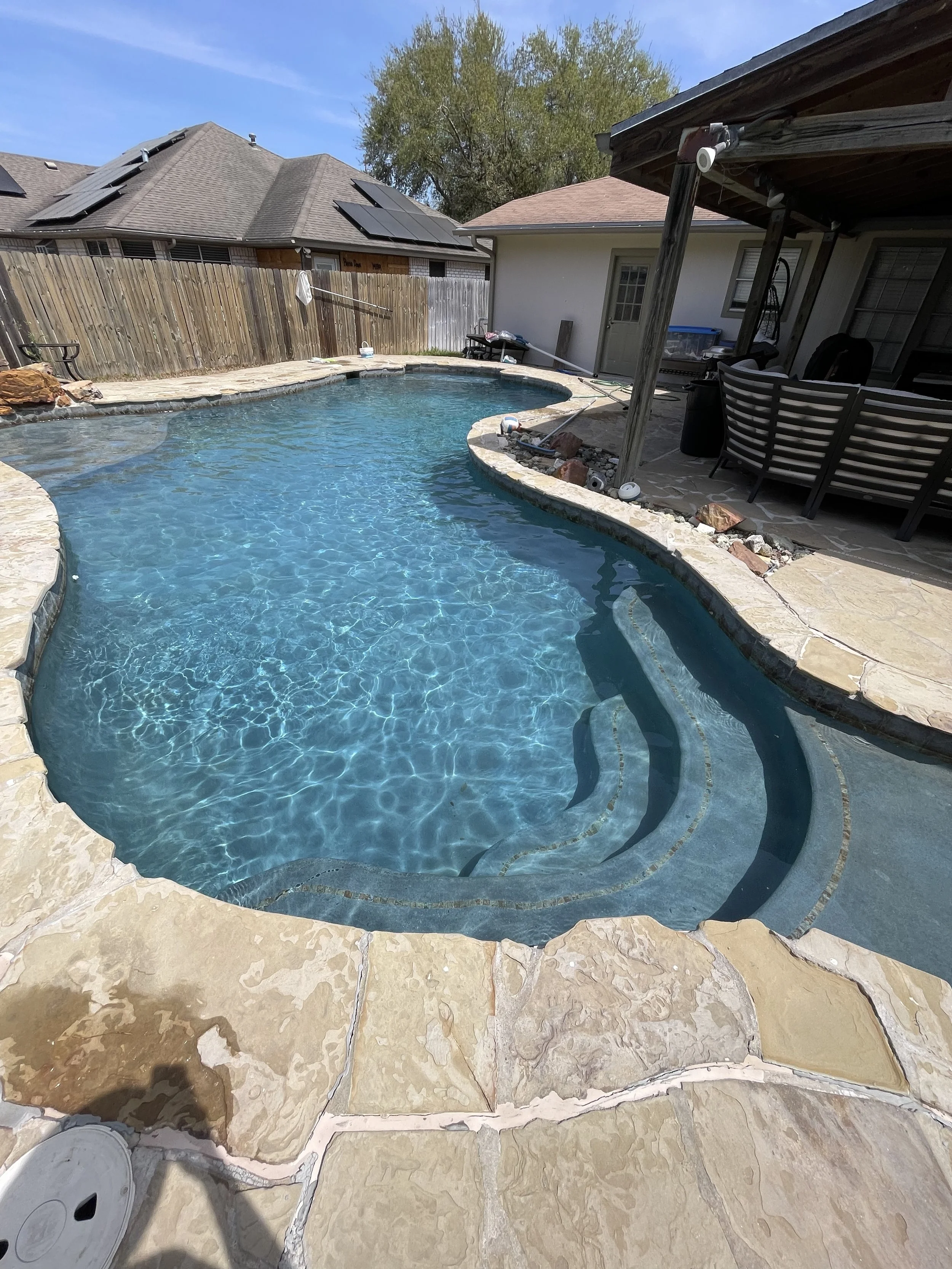 A backyard swimming pool with stone edging, clear blue water, and built-in steps. Surrounding the pool is a stone deck, with a covered patio area featuring outdoor furniture. Wooden fences and neighboring houses with solar panels are visible in the background.