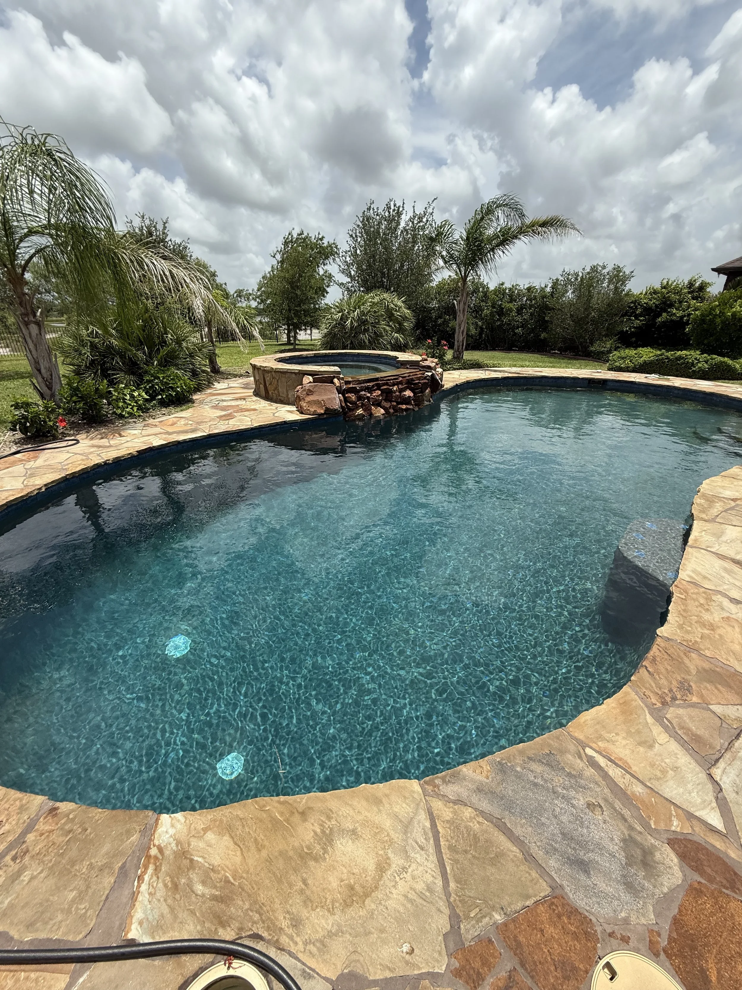 Shot of a backyard with a swimming pool, hot tub, and palm trees under a partly cloudy sky.