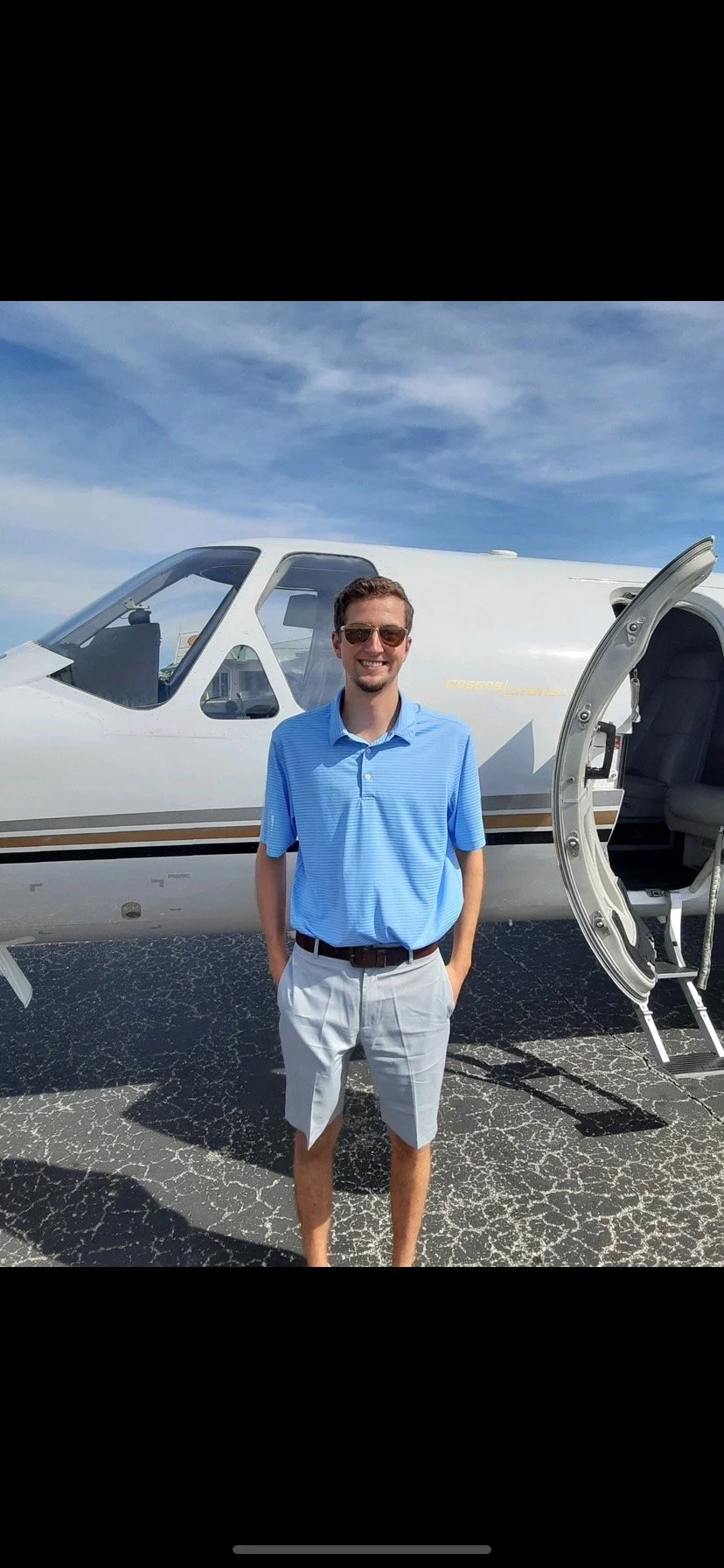 A man in sunglasses wearing a blue polo shirt and white shorts standing in front of a private jet on an airport tarmac with a partly cloudy sky.