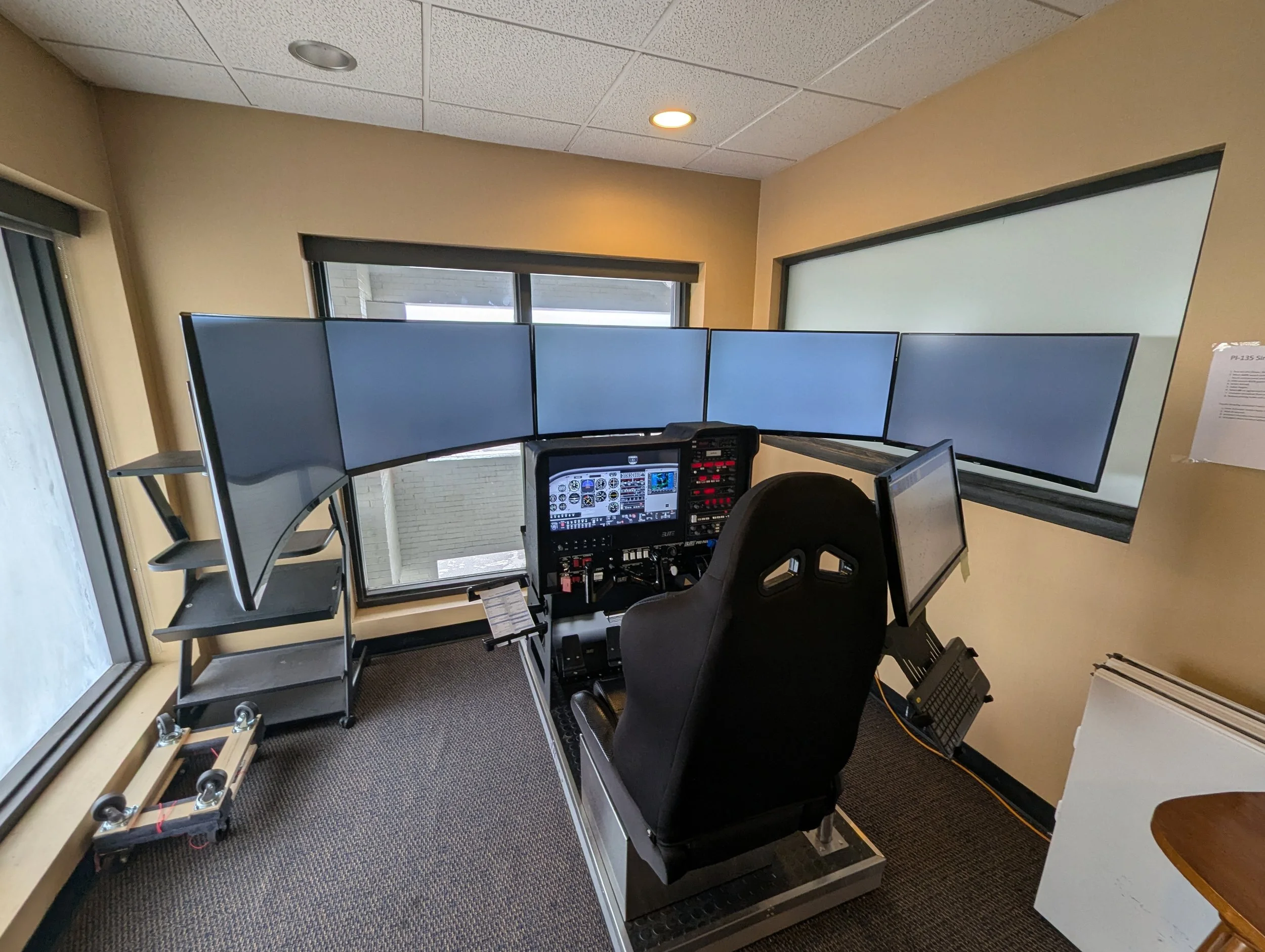 A flight simulator cockpit with five large screens, a pilot seat, and flight instruments in an office-like room with windows and beige walls.