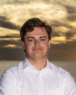 A young man in a white shirt outdoors during sunset with a cloudy sky in the background.