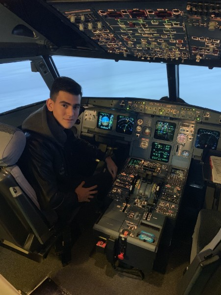 Young man sitting in the cockpit of an airplane simulator surrounded by flight controls and instruments, with a view of the sky outside the windshield.