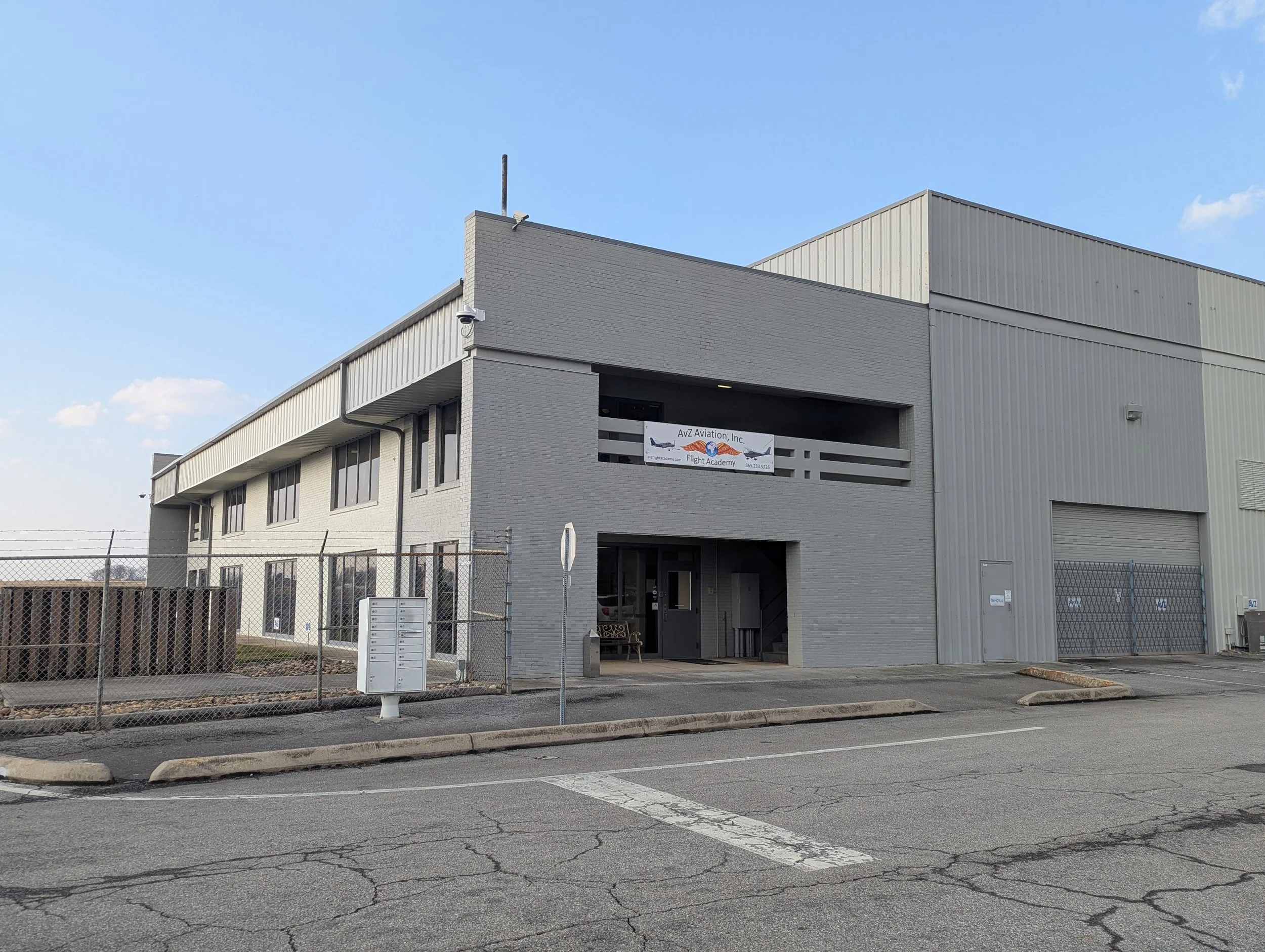An exterior photo of AvZ's office at McGhee Tyson Airport, which is located on the second floor of the building. Attached to the right of the building is a large community hangar, and to the left is a chain link fence that surrounds the airport ramp.