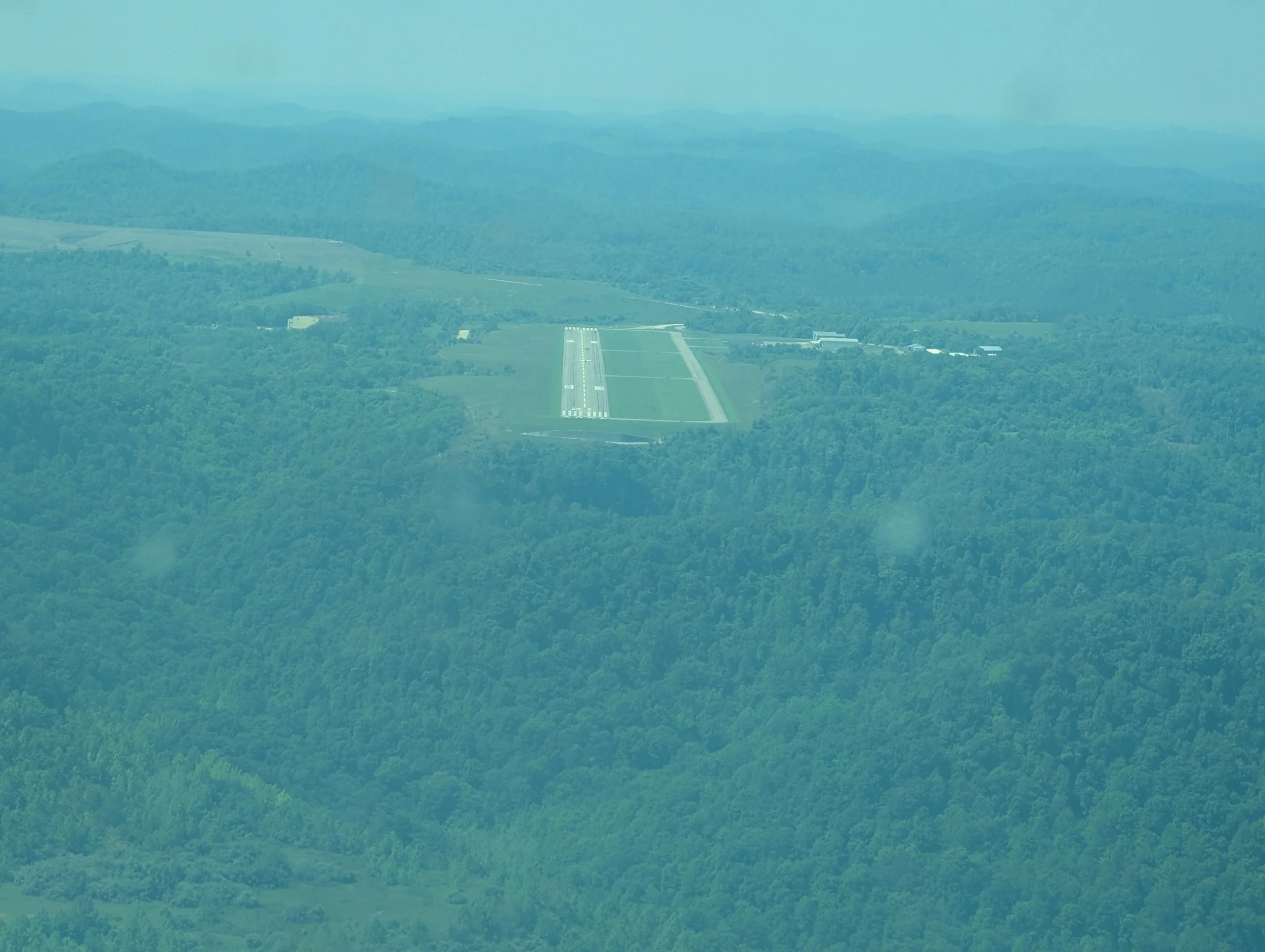 An aerial view of a the runways at the KSJS airport in Kentucky, surrounded by lush green forest with rolling hills in the background. This airport is a popular cross country destination for our students due to a restaurant being located at the field