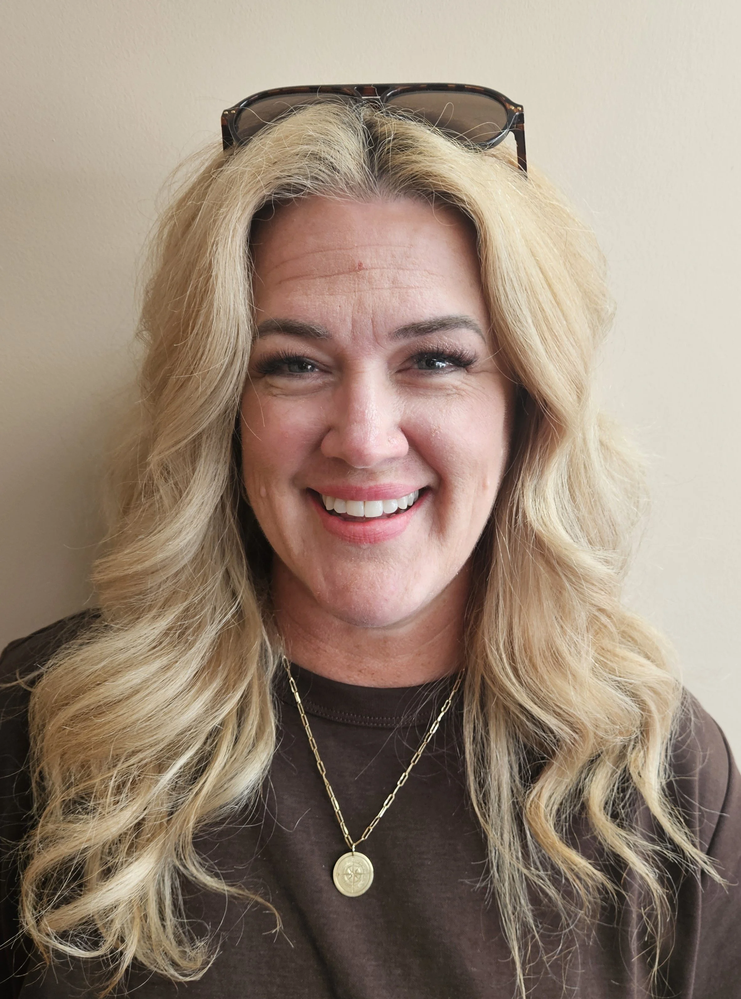 A woman with long blonde hair, wearing glasses on her head, smiling in front of a beige wall.